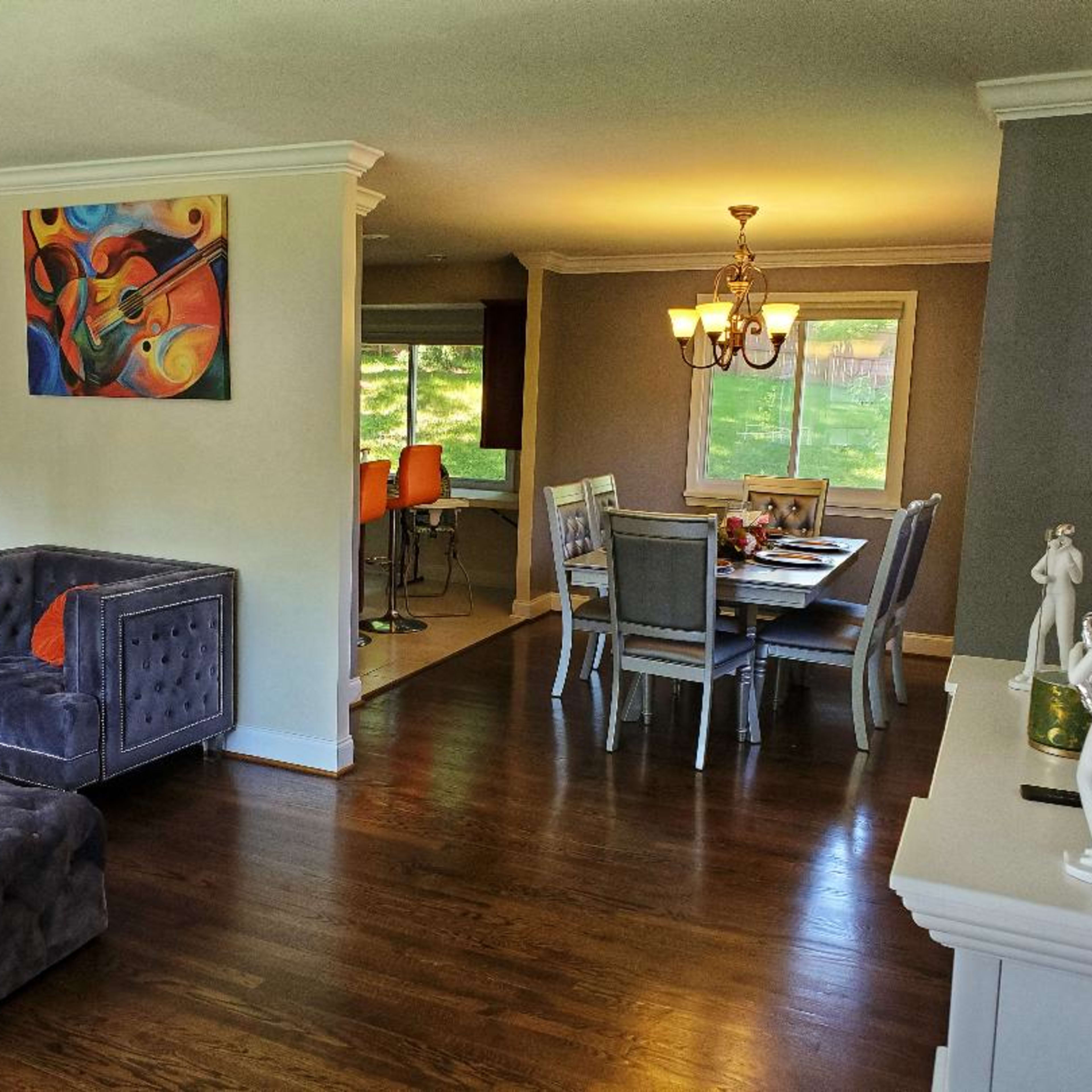 A well-lit living area with a tufted gray sofa, a dining table with chairs, and a staircase leading to an upper level.