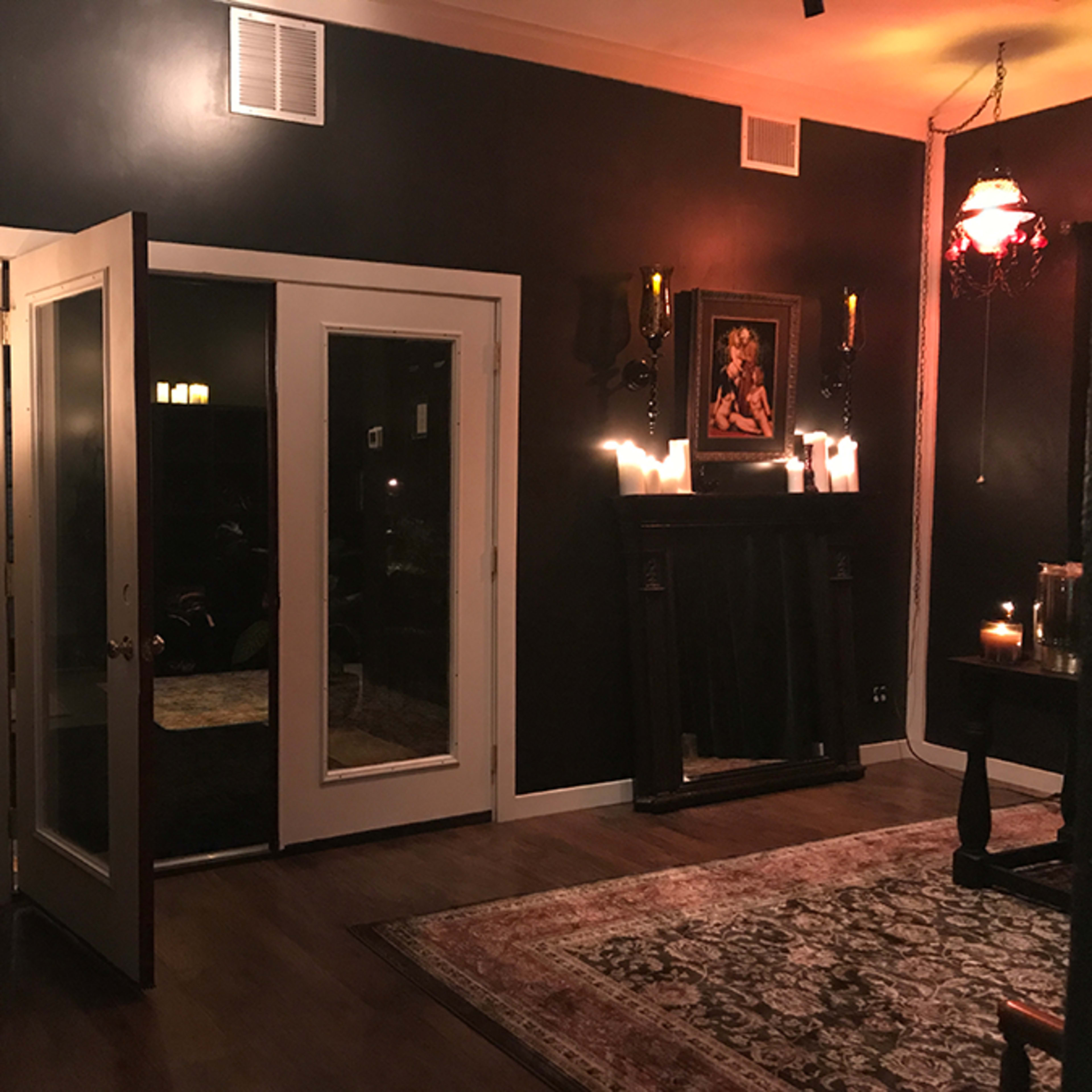 A dimly lit room features a pair of open double doors, a dark-painted wall, a fireplace with candles, and a large rug on the wooden floor.