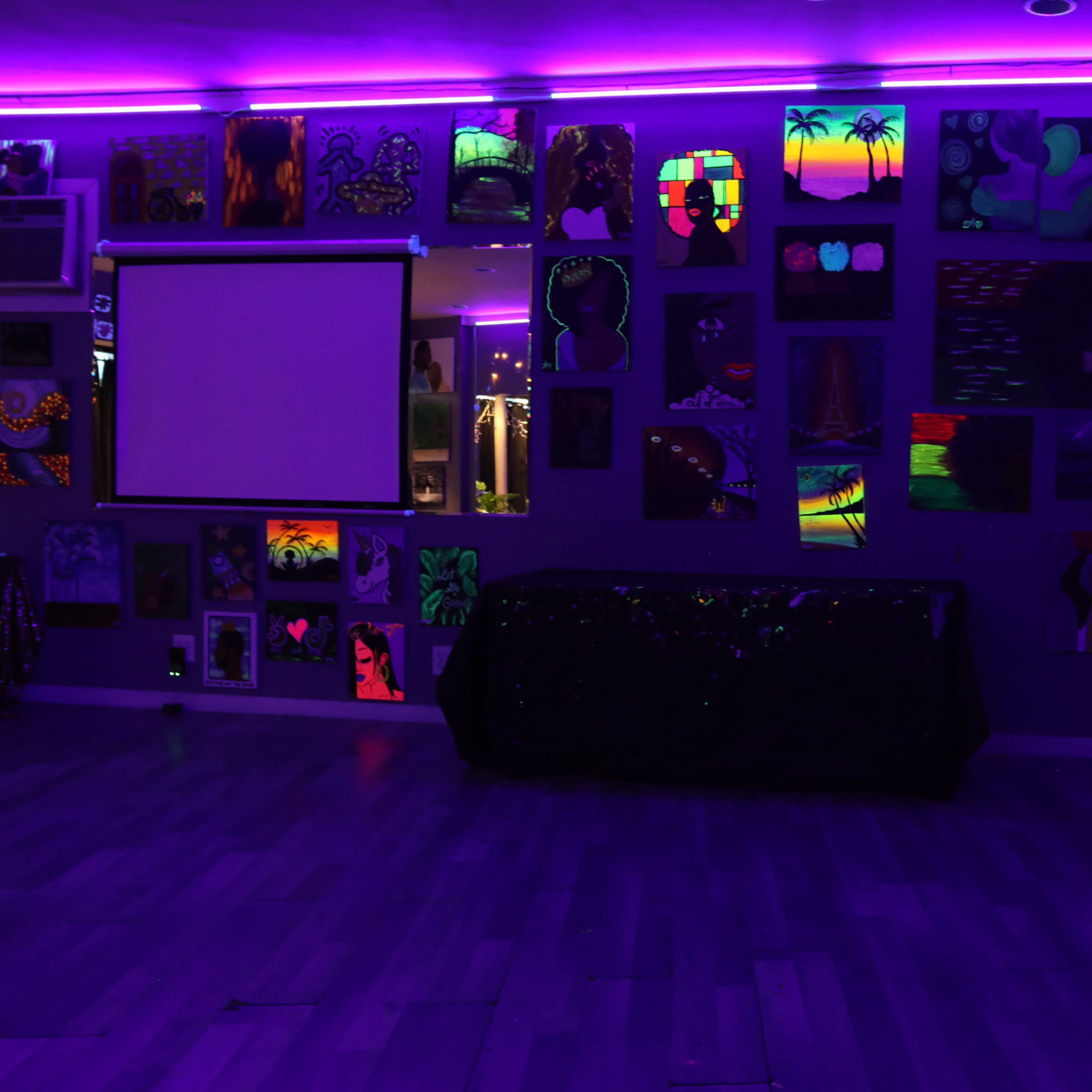 A dimly lit room with purple neon lights highlighting a wall covered in various colorful artworks, accompanied by a projector screen and a dark table in the center.