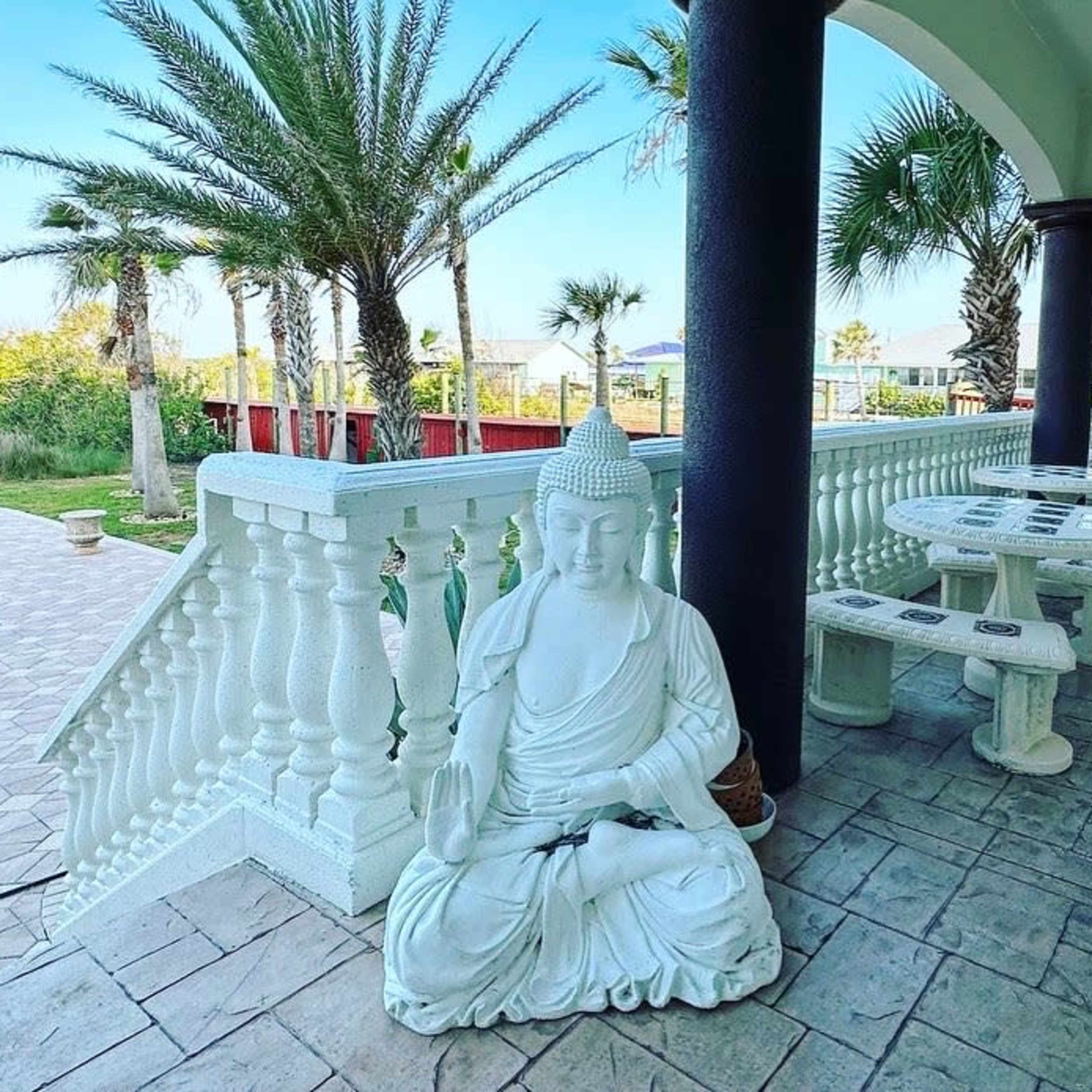 A white Buddha statue sits on the patio beside stone tables and chairs, with palm trees visible in the background.