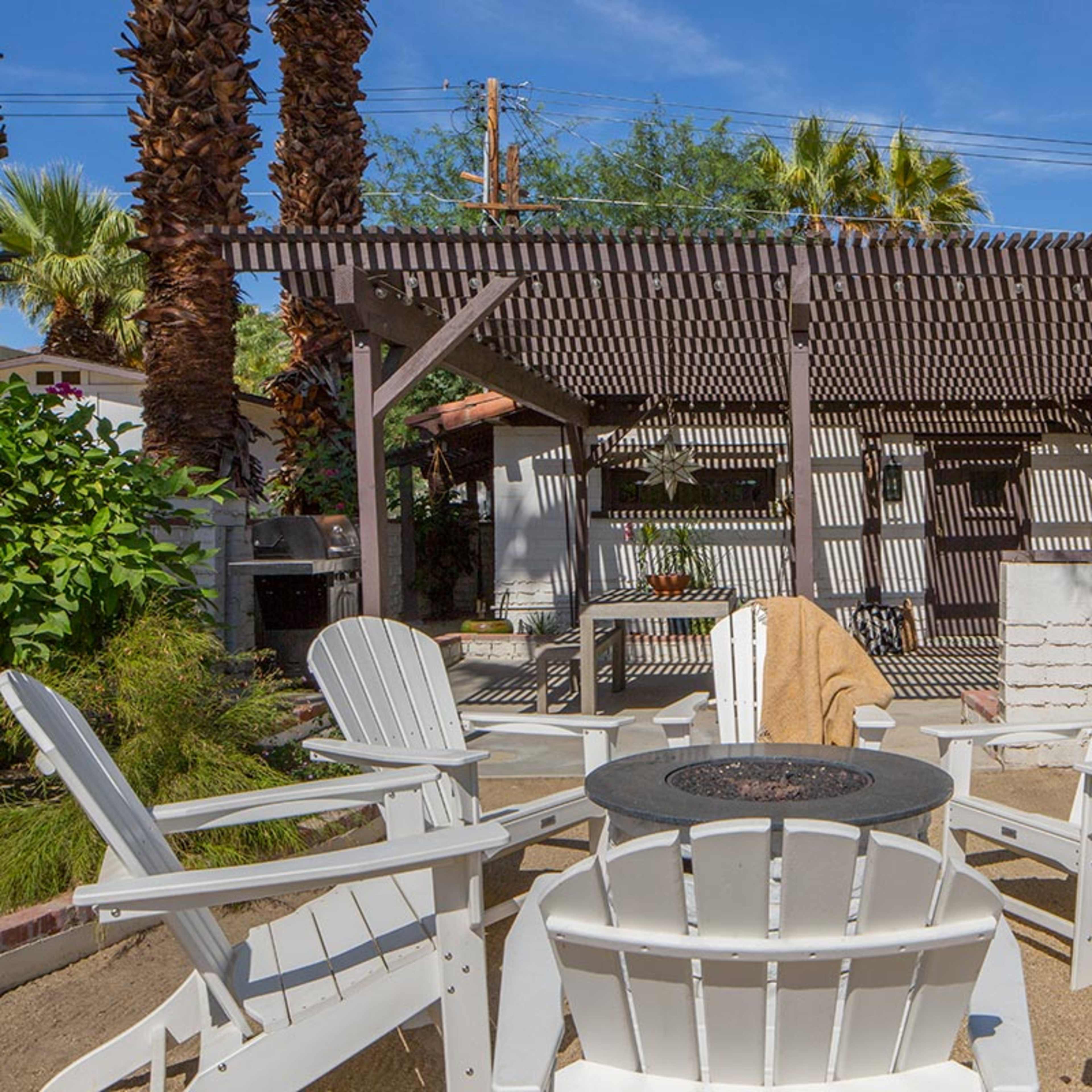 A shaded outdoor seating area features white Adirondack chairs arranged around a circular fire pit in front of a pergola and landscaped greenery.