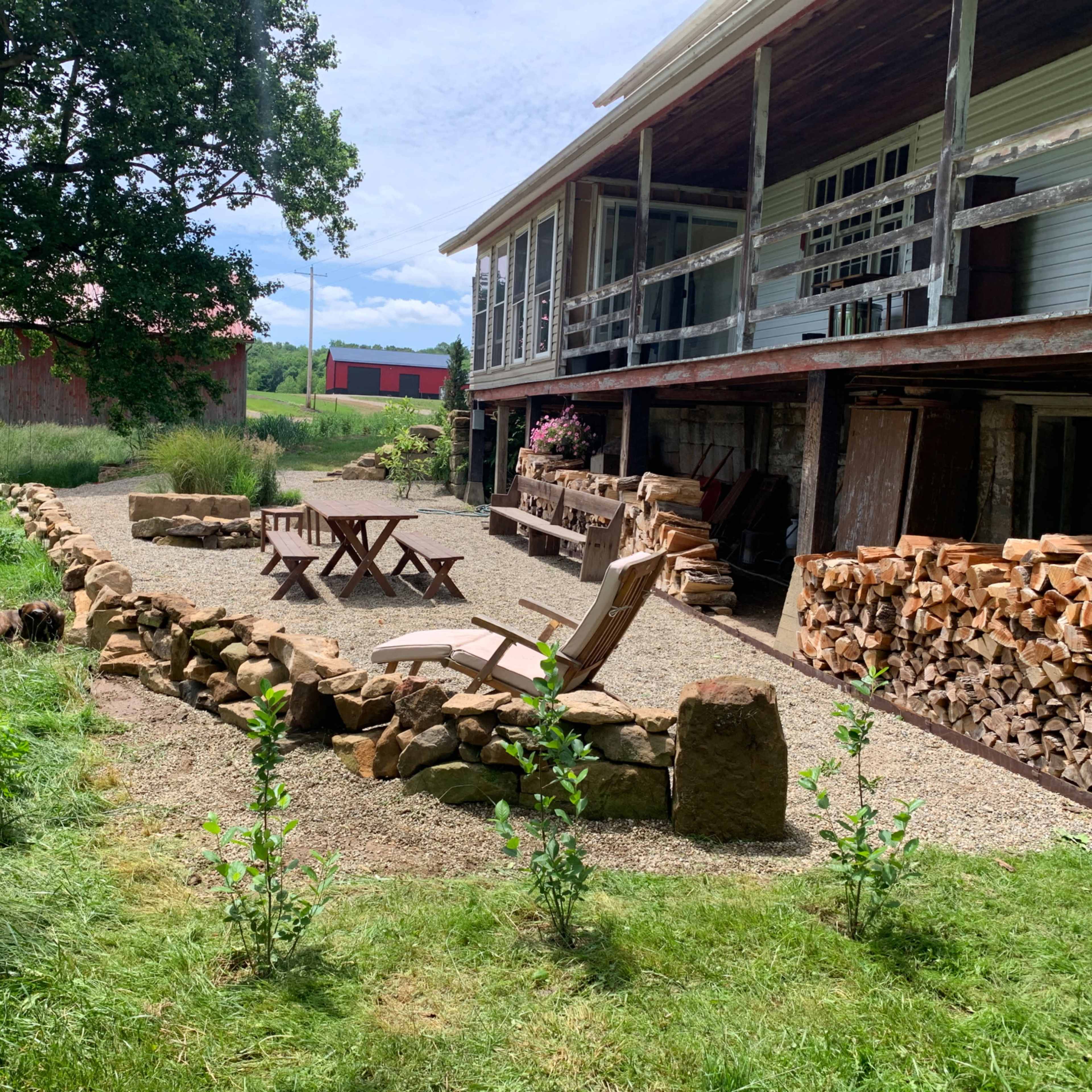 A gravel patio area with wooden seating and stacked firewood, adjacent to a multi-level house surrounded by greenery.