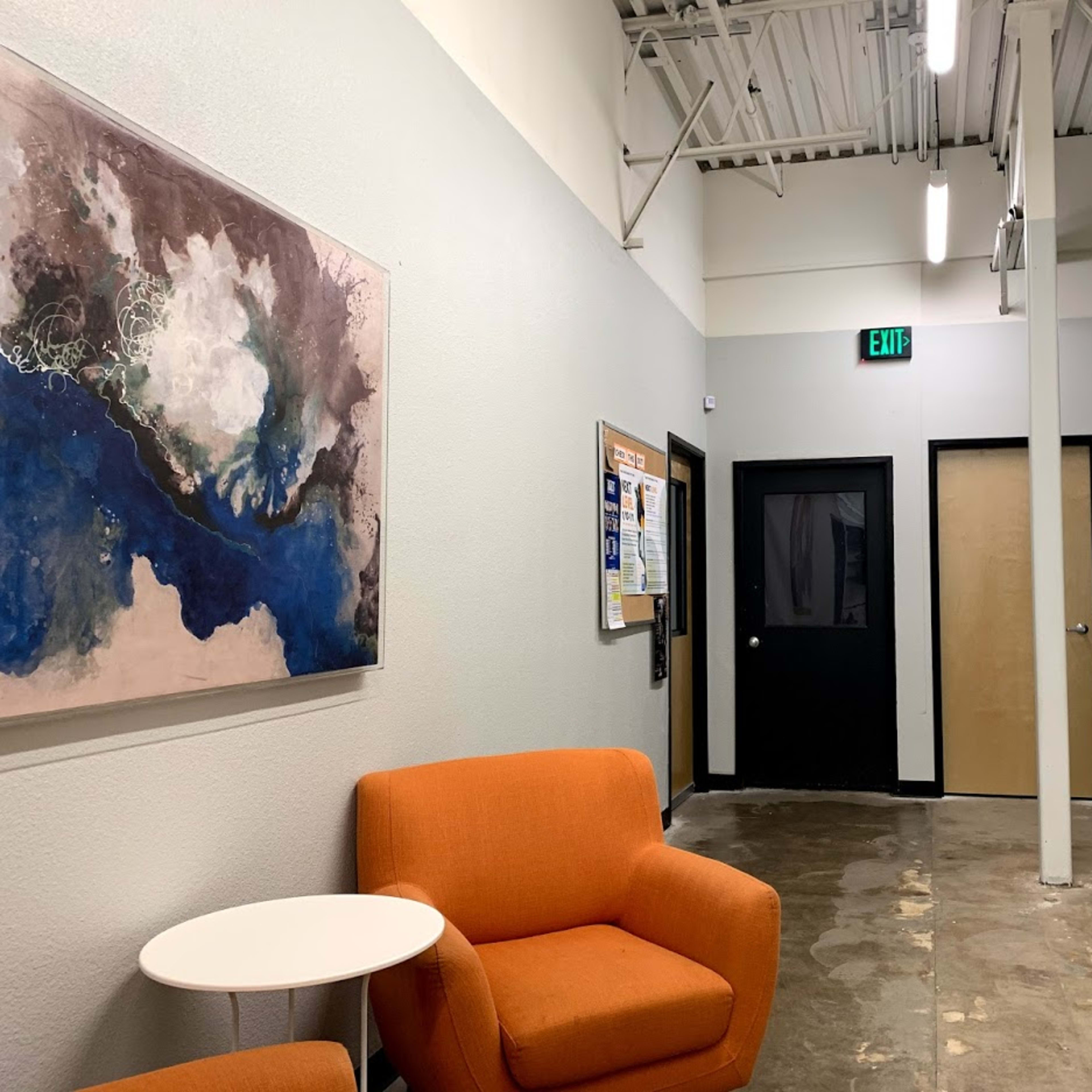 A hallway with a seating area featuring two orange chairs, an abstract painting on the wall, and doorways leading to other rooms.