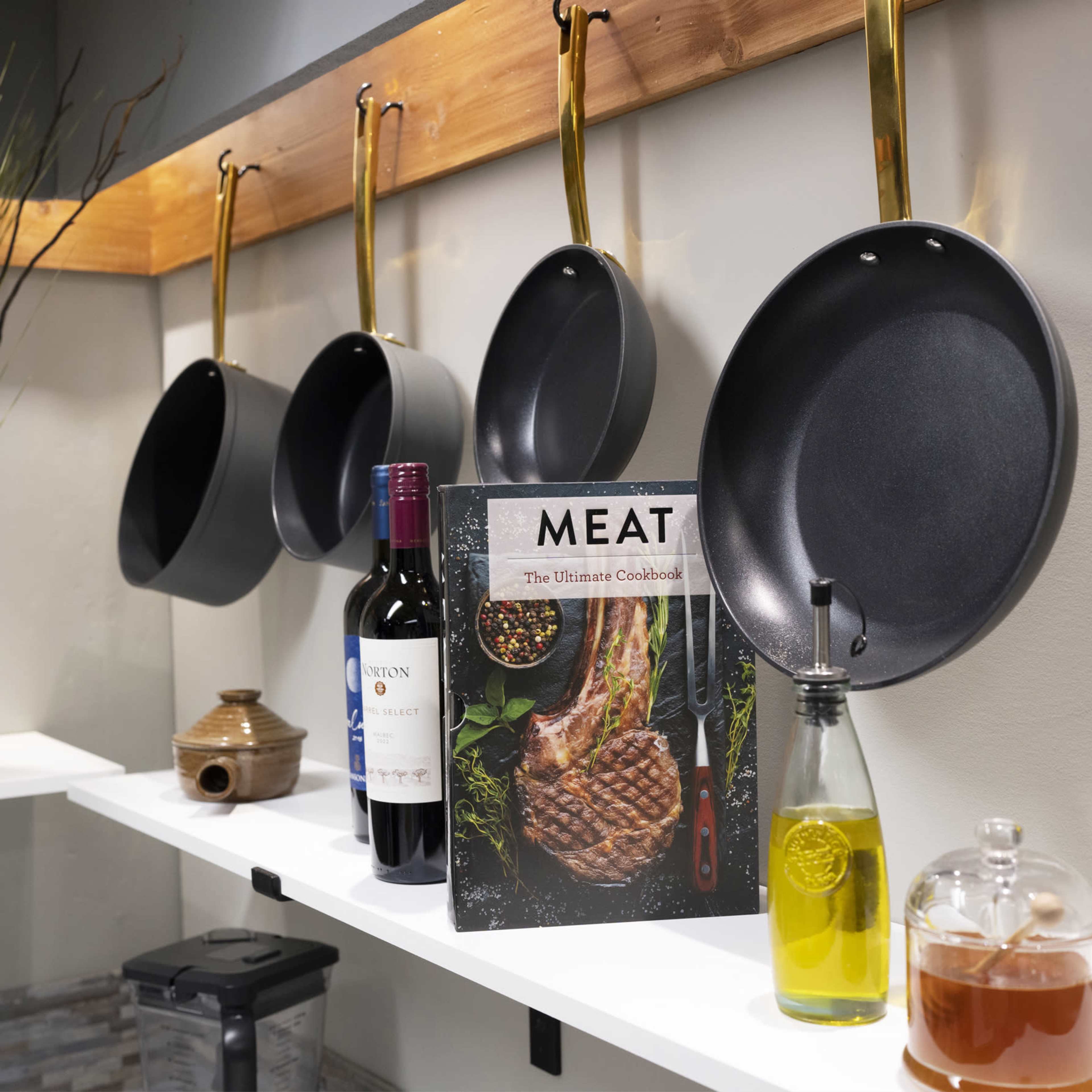 A kitchen countertop displays hanging pots, a bottle of red wine, an oil bottle, a cookbook titled "MEAT," and a small honey jar.