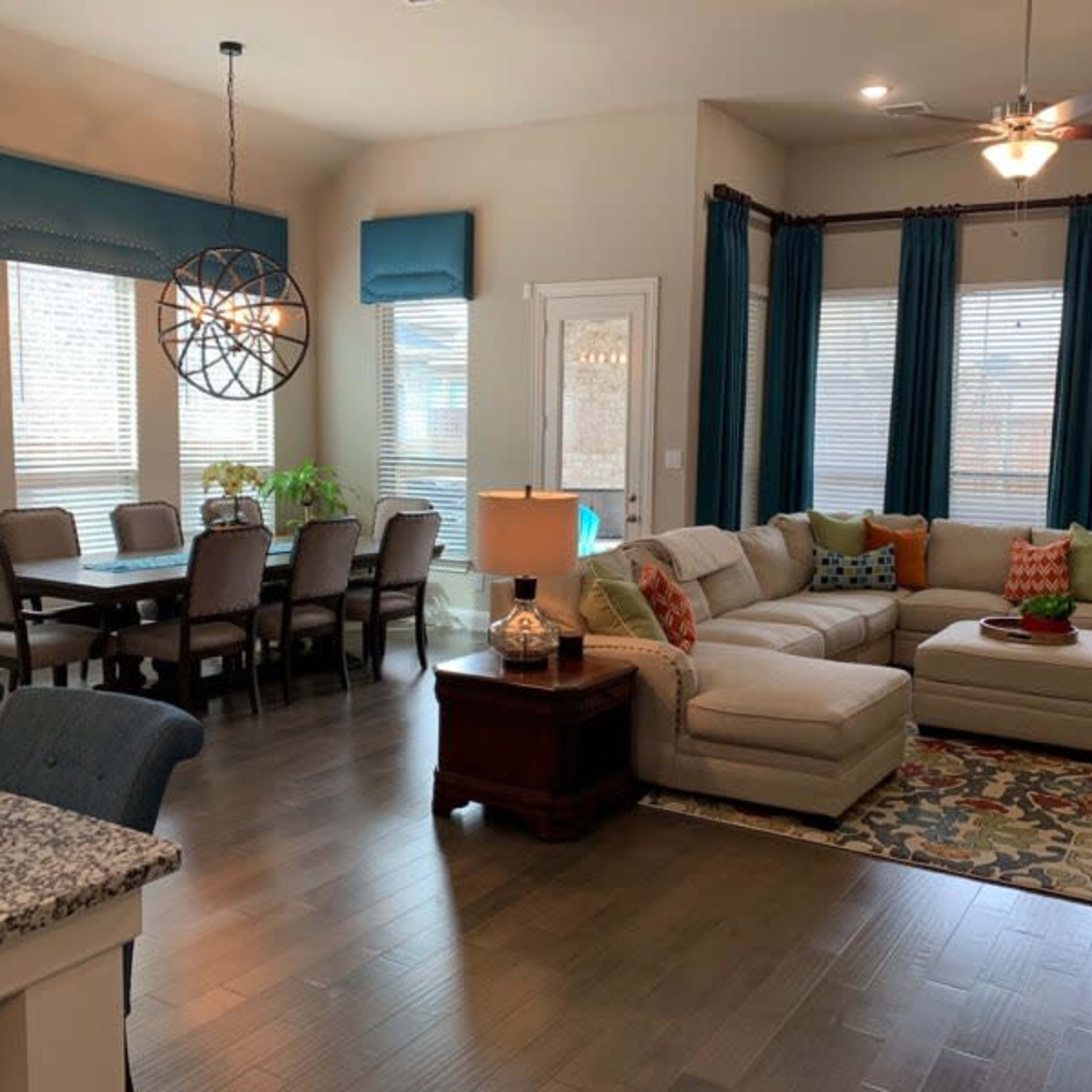 A living room and dining area featuring a sectional sofa, a dining table with chairs, and large windows with blue curtains.