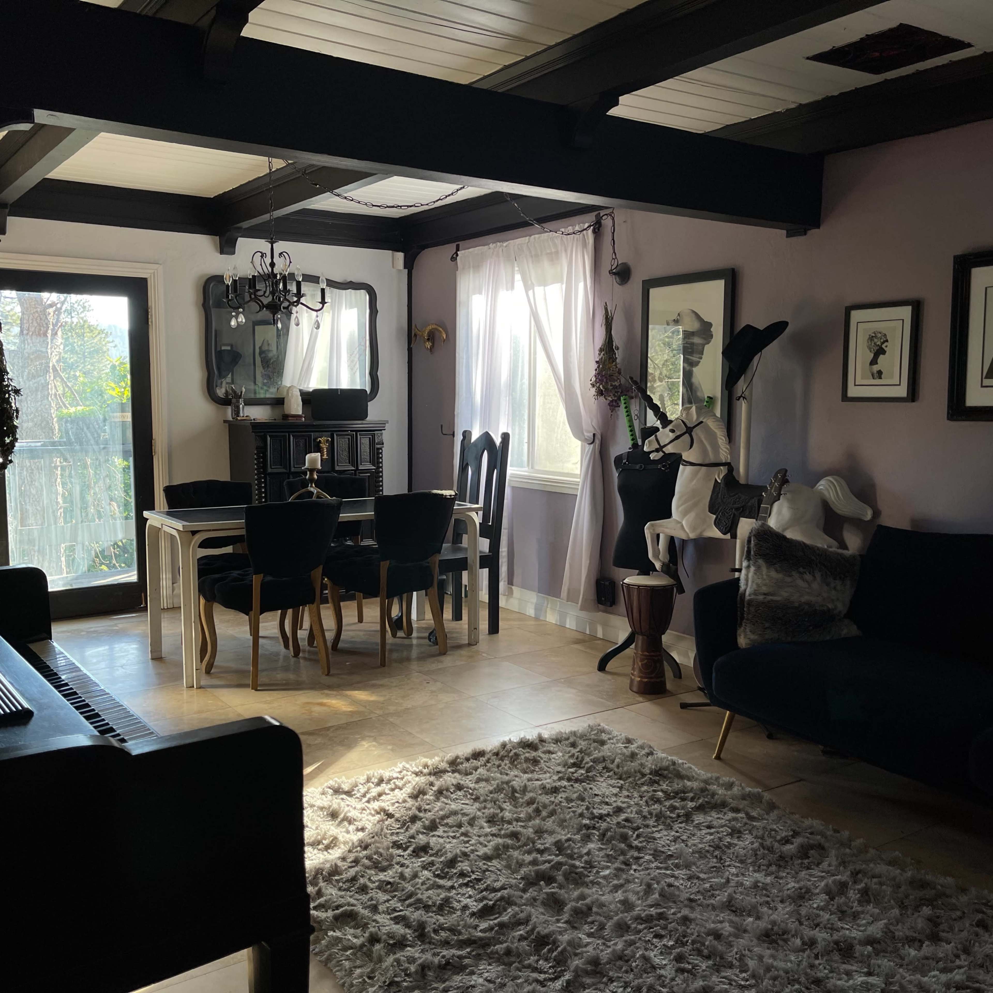 A living room featuring a dining area with a white table and black chairs, a dark sofa, large windows with sheer curtains, and various framed artworks on the walls.