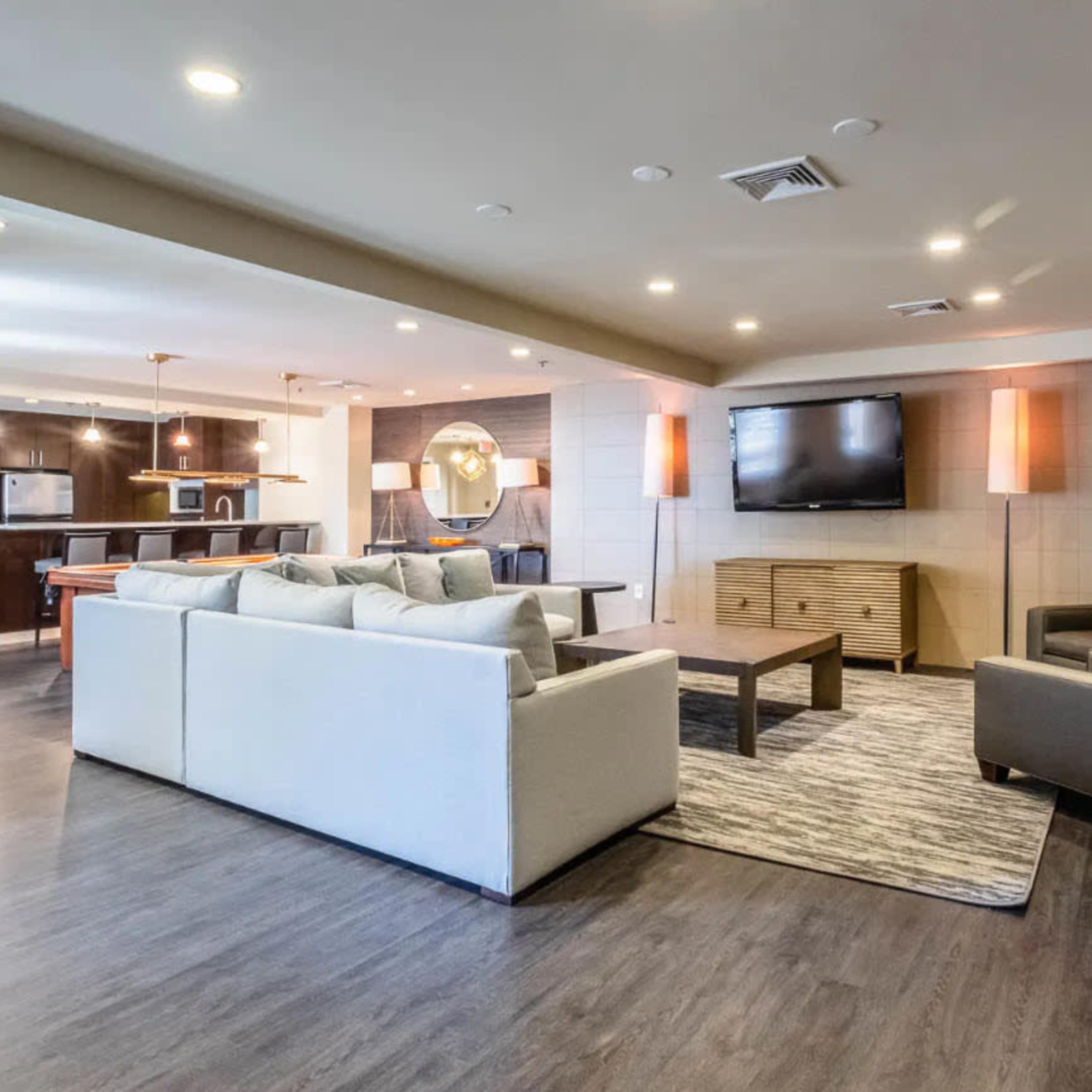 A modern living area with a sofa, two armchairs, a coffee table, and a wall-mounted television, leading into a kitchen space in the background.