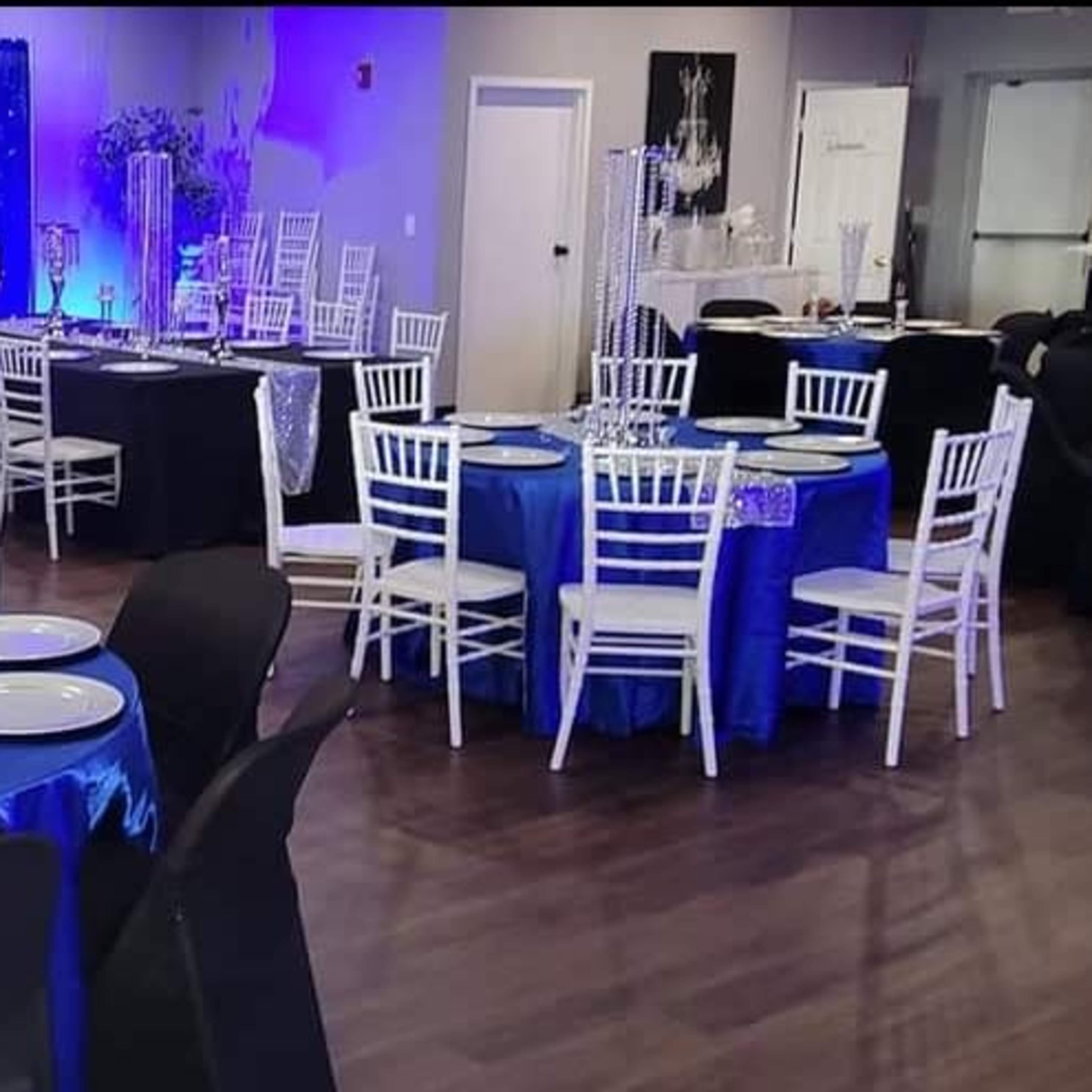 A banquet hall arranged with round tables covered in blue tablecloths and black chairs, featuring decorative centerpieces and a backdrop with purple lighting.