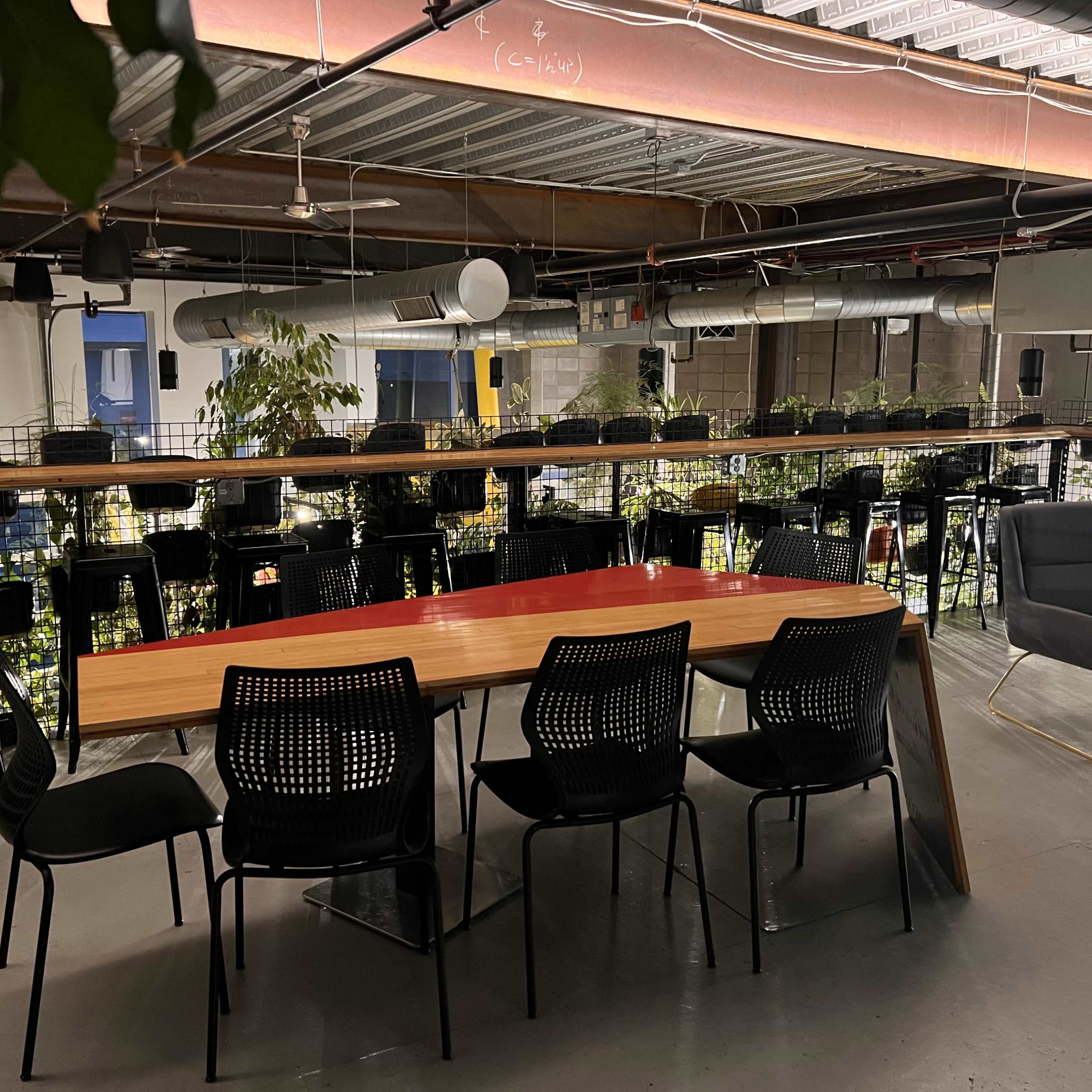 A modern indoor workspace featuring a long wooden table surrounded by black chairs, with plants and high-top seating areas in the background.
