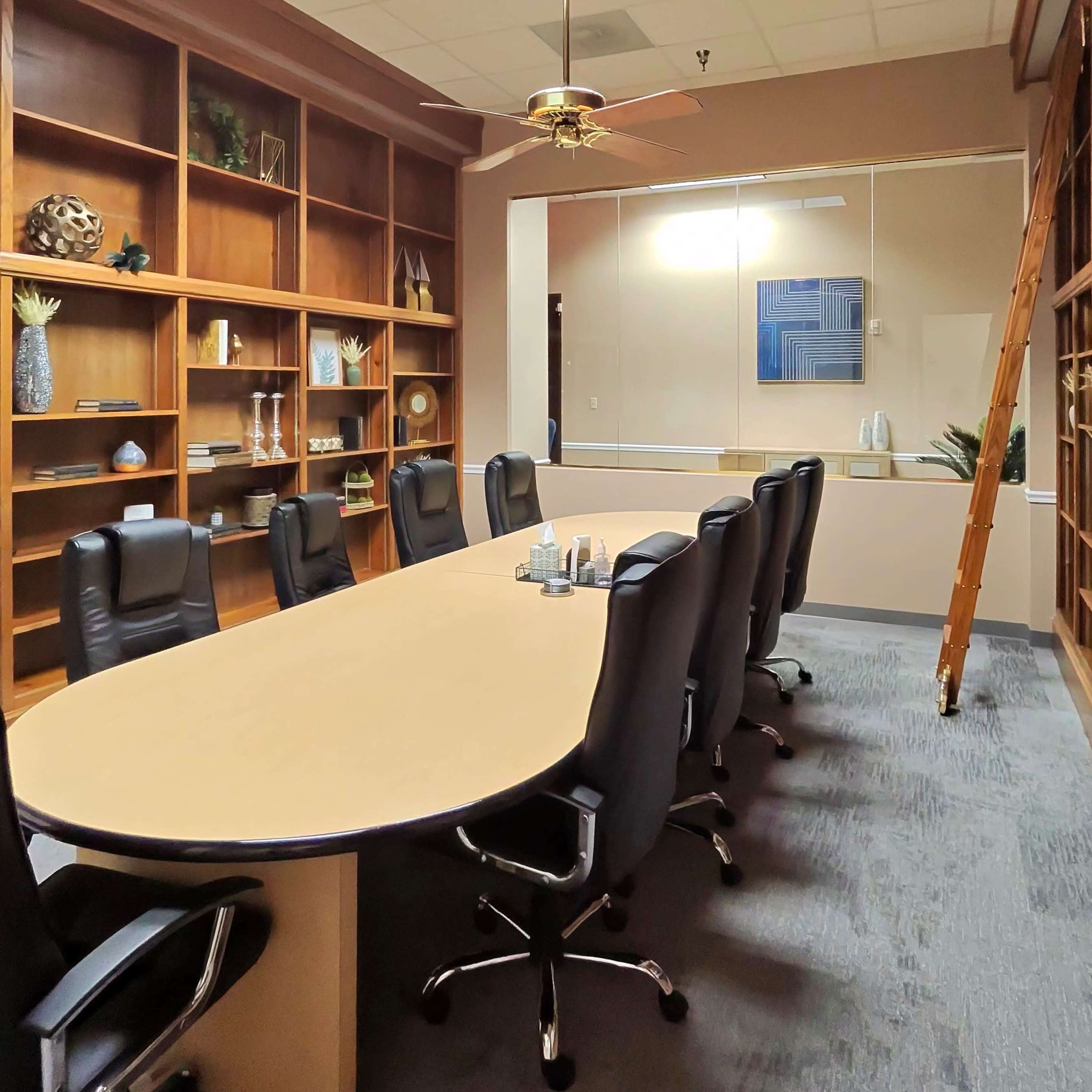 A conference room features a long oval table surrounded by black chairs, with built-in shelves displaying decorative items and a ladder against the wall.