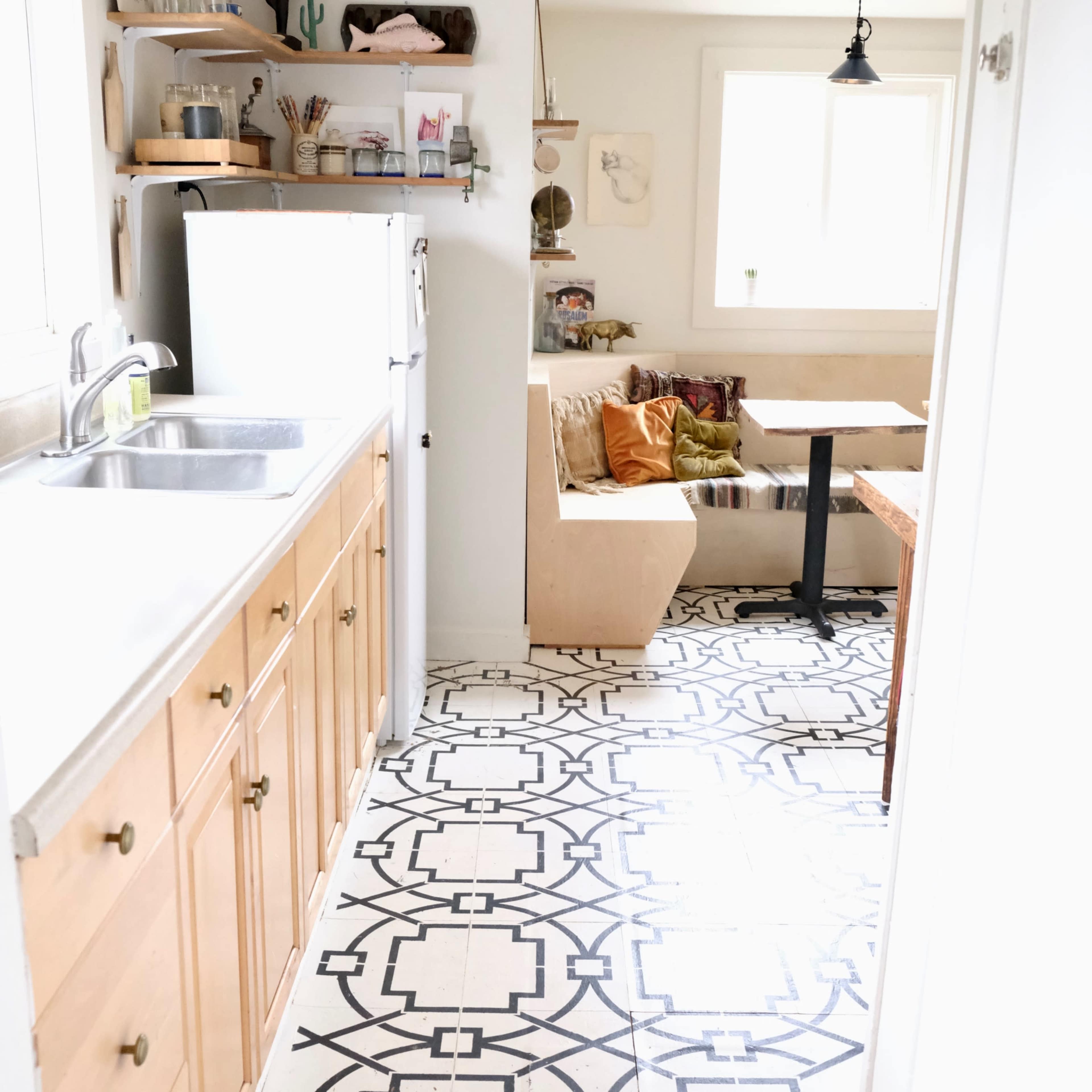 A narrow kitchen features wooden cabinets, a stainless steel sink, and patterned tiles leading to a dining area with a built-in bench and a table.