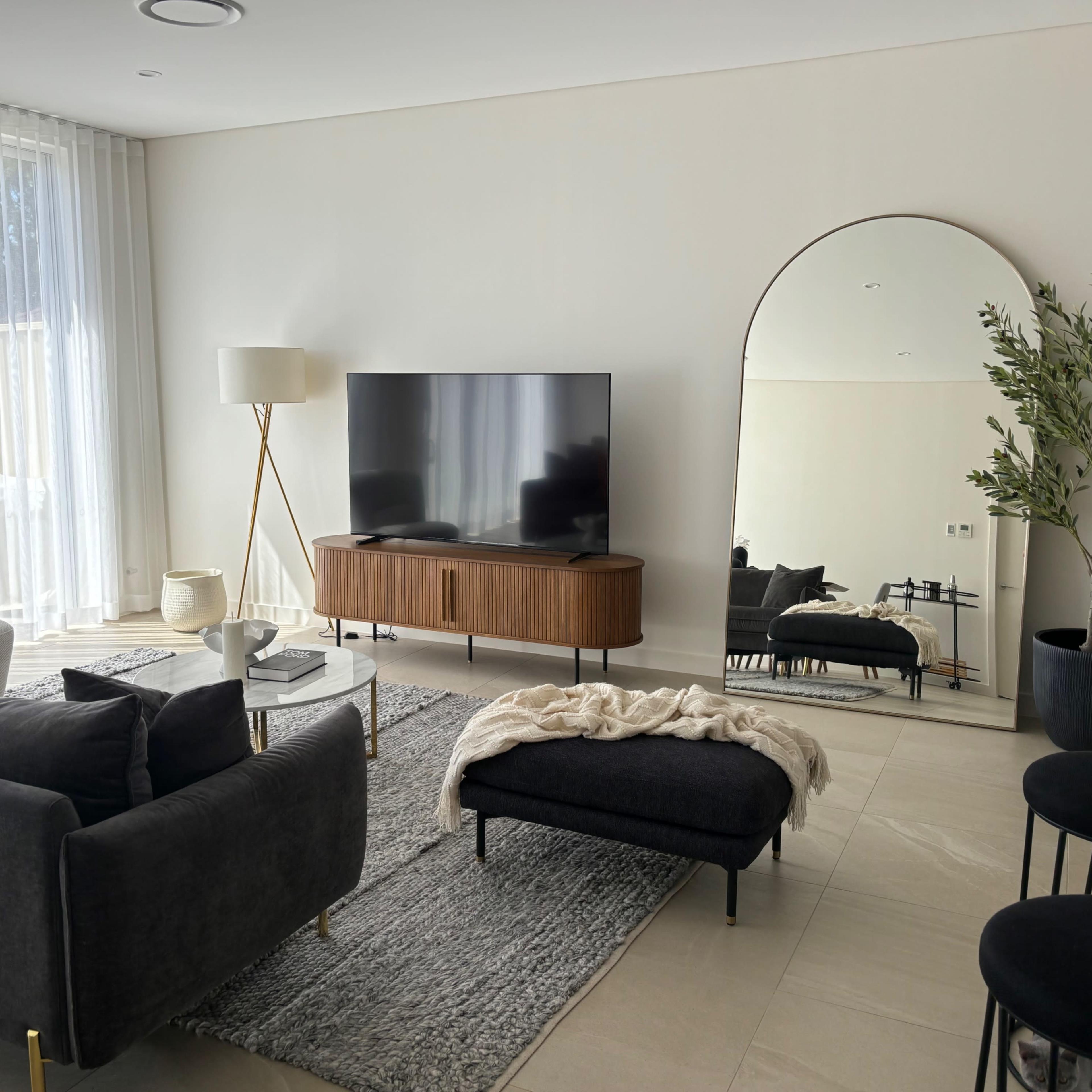 The living room features a dark sofa, a wooden TV cabinet, a large mirror, and a contemporary lamp, with light streaming through sheer curtains.