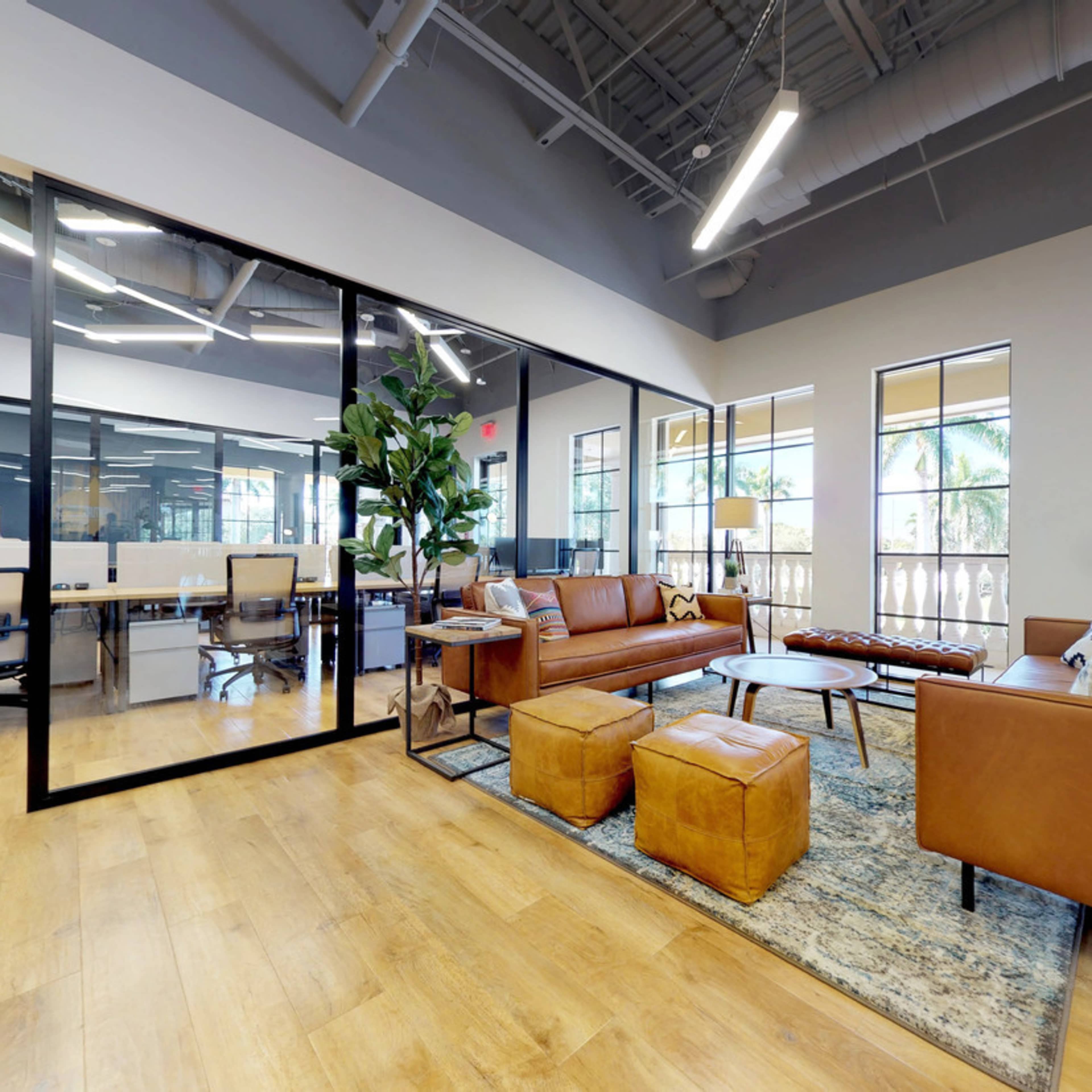 A modern office space featuring a lounge area with a brown leather couch, coffee table, and potted plant, adjacent to a workspace with desks and glass partitions.