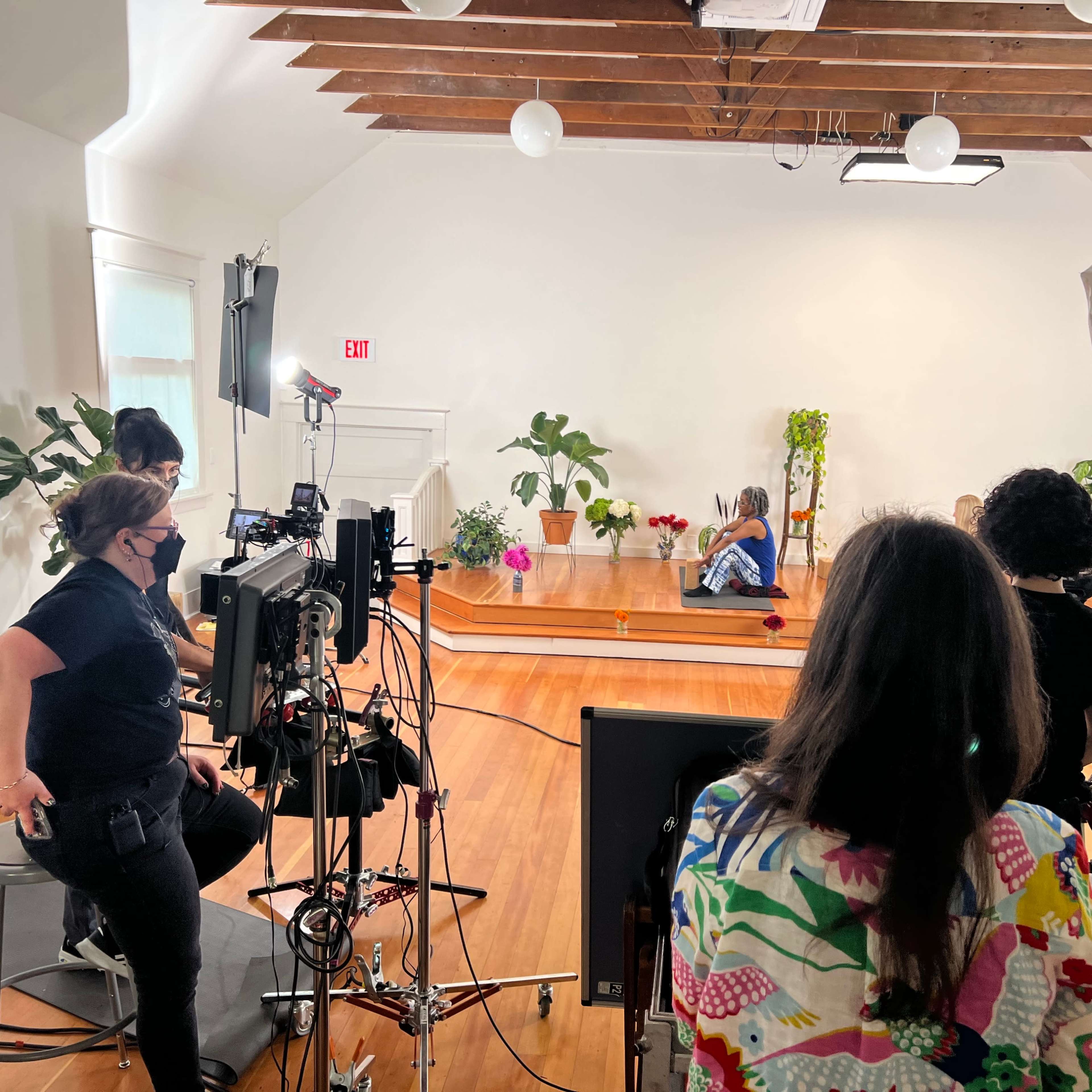 A filming setup in a bright room with wooden floors, where a crew is working around a person seated on a stage surrounded by plants and lighting equipment.