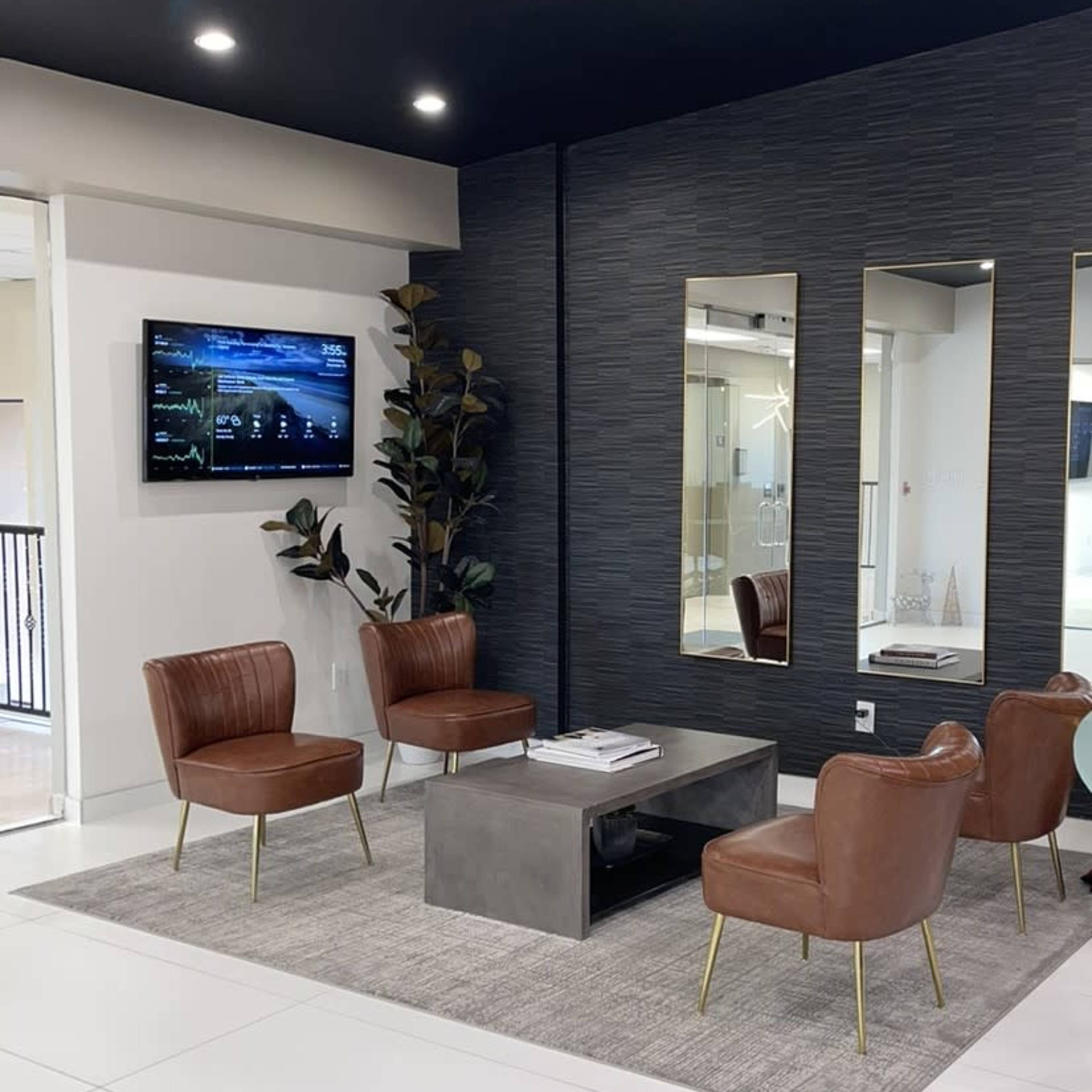 A modern waiting area featuring four brown leather chairs arranged around a metal table on a light gray carpet, with a television mounted on the wall and two mirrors flanking it.