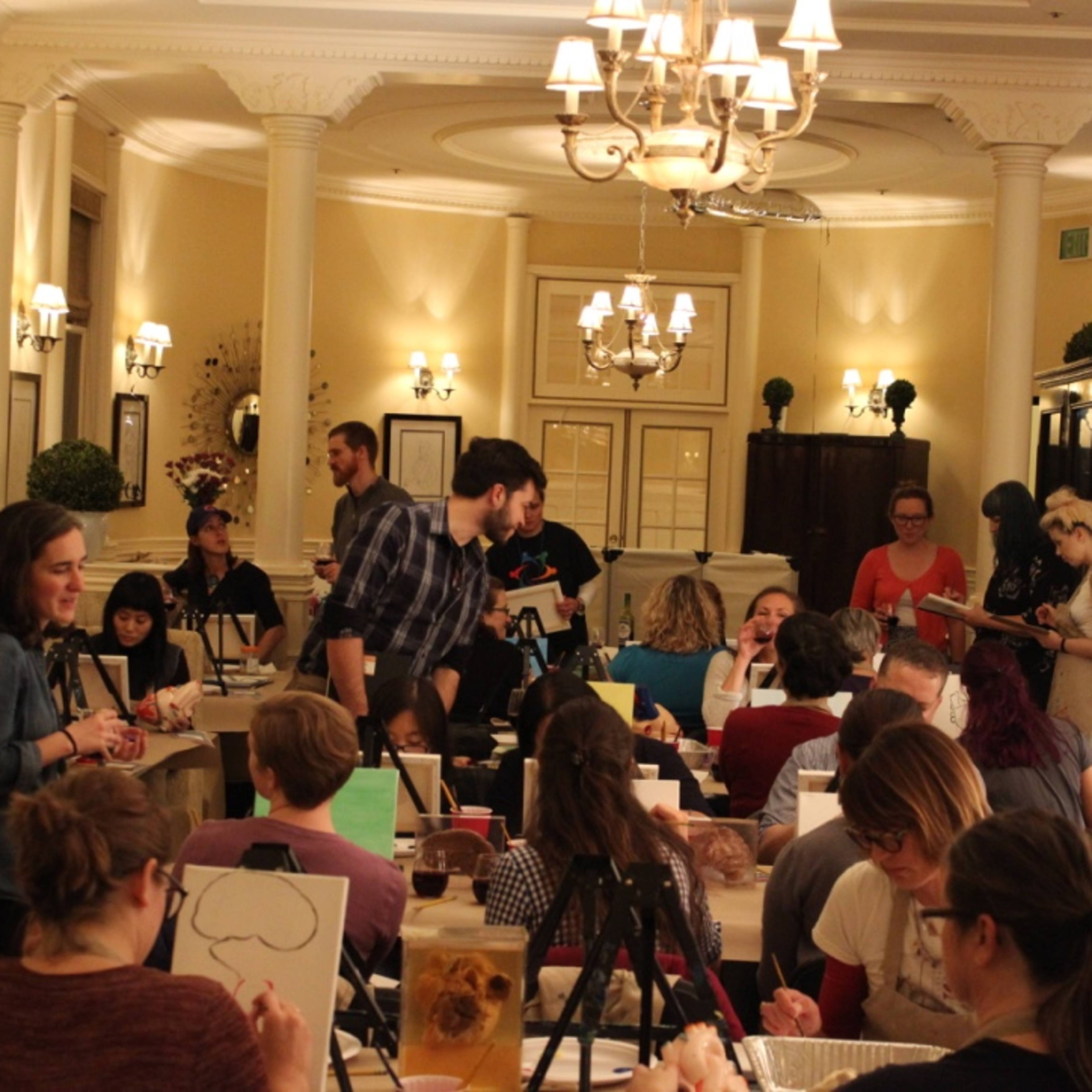 A group of people participates in a painting class in an elegantly decorated room with tables, easels, and art supplies.