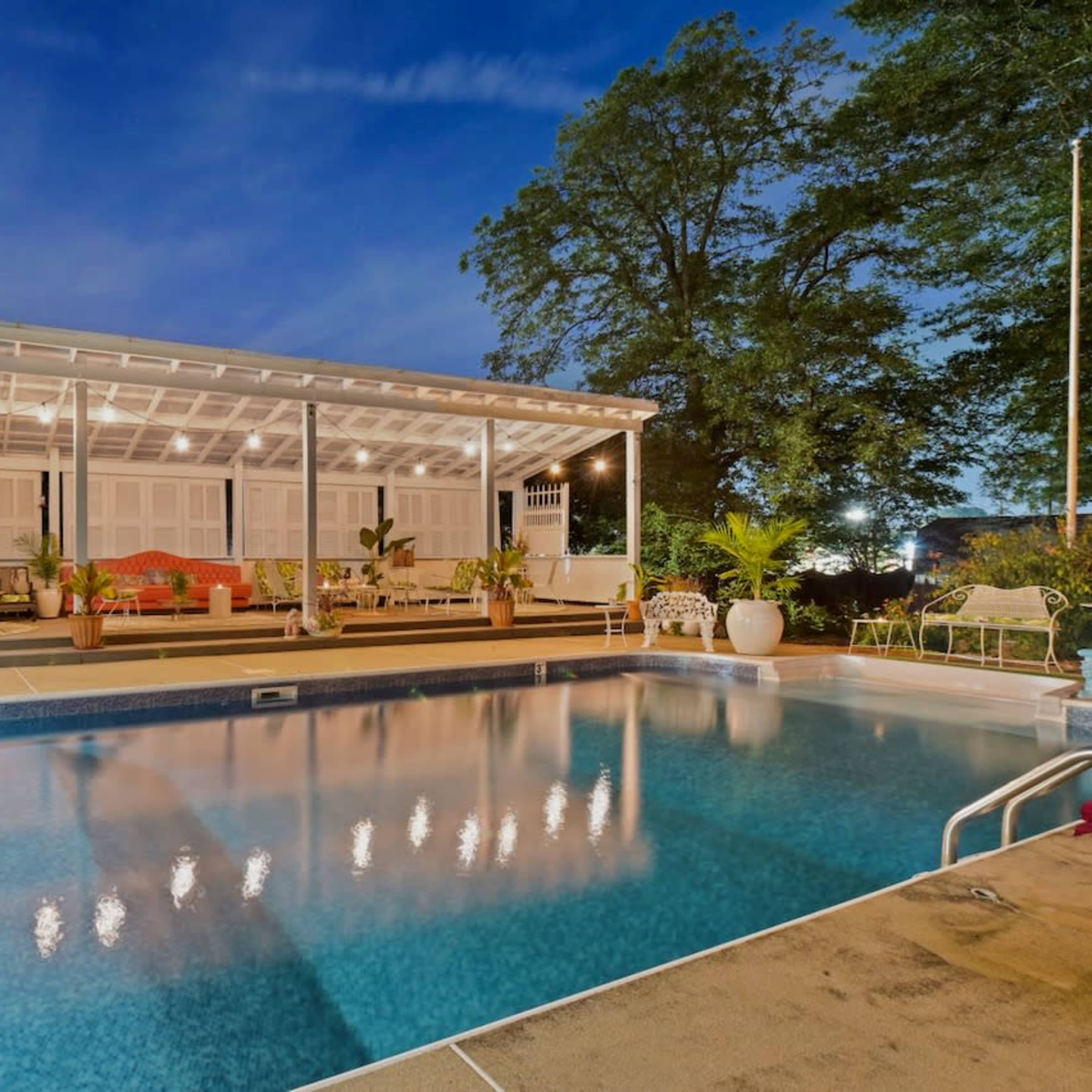 A modern backyard features a pool in front of a covered seating area with lush greenery at dusk.