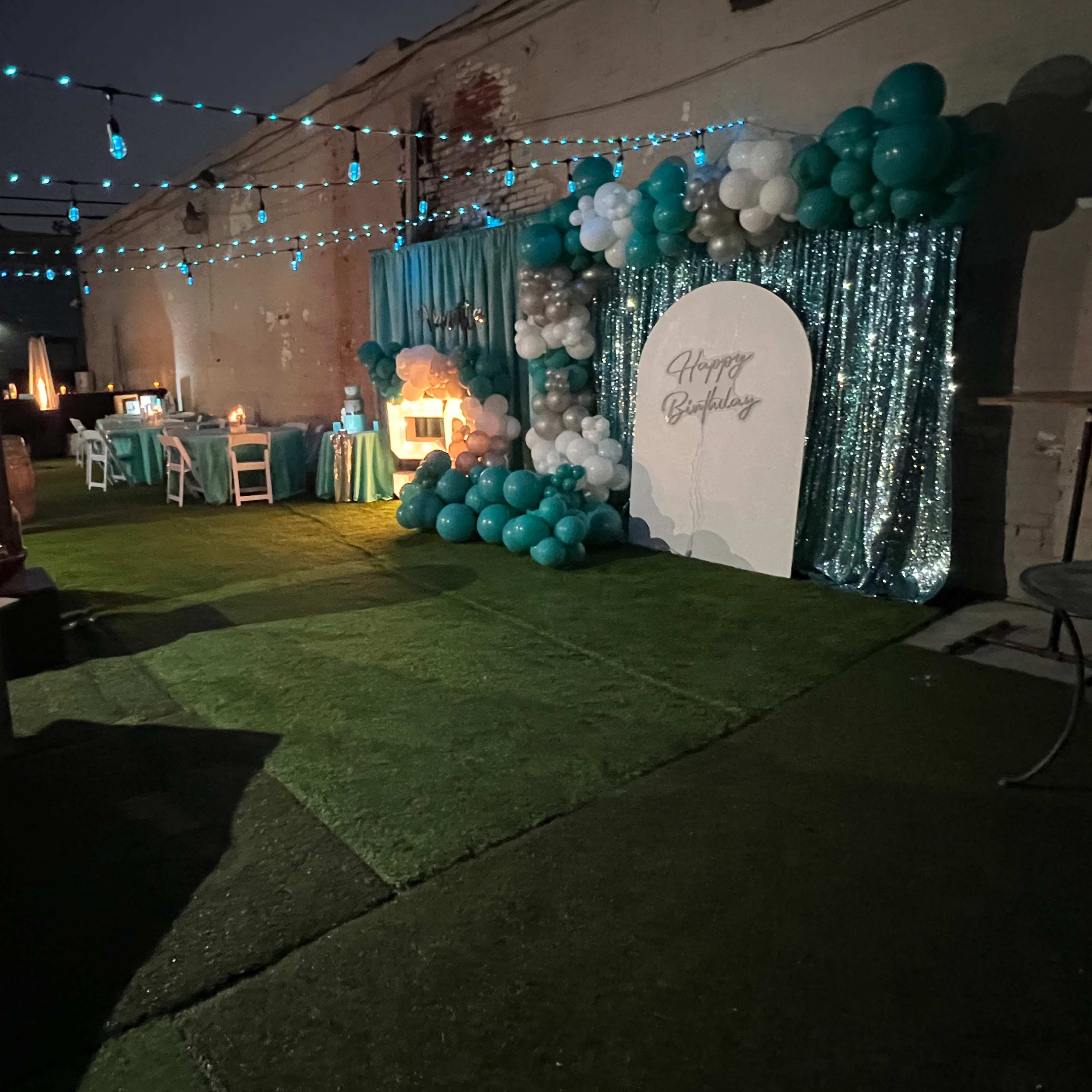 A birthday party setup outdoors, featuring a decorated backdrop with balloons, a table set for guests, and a fire pit.