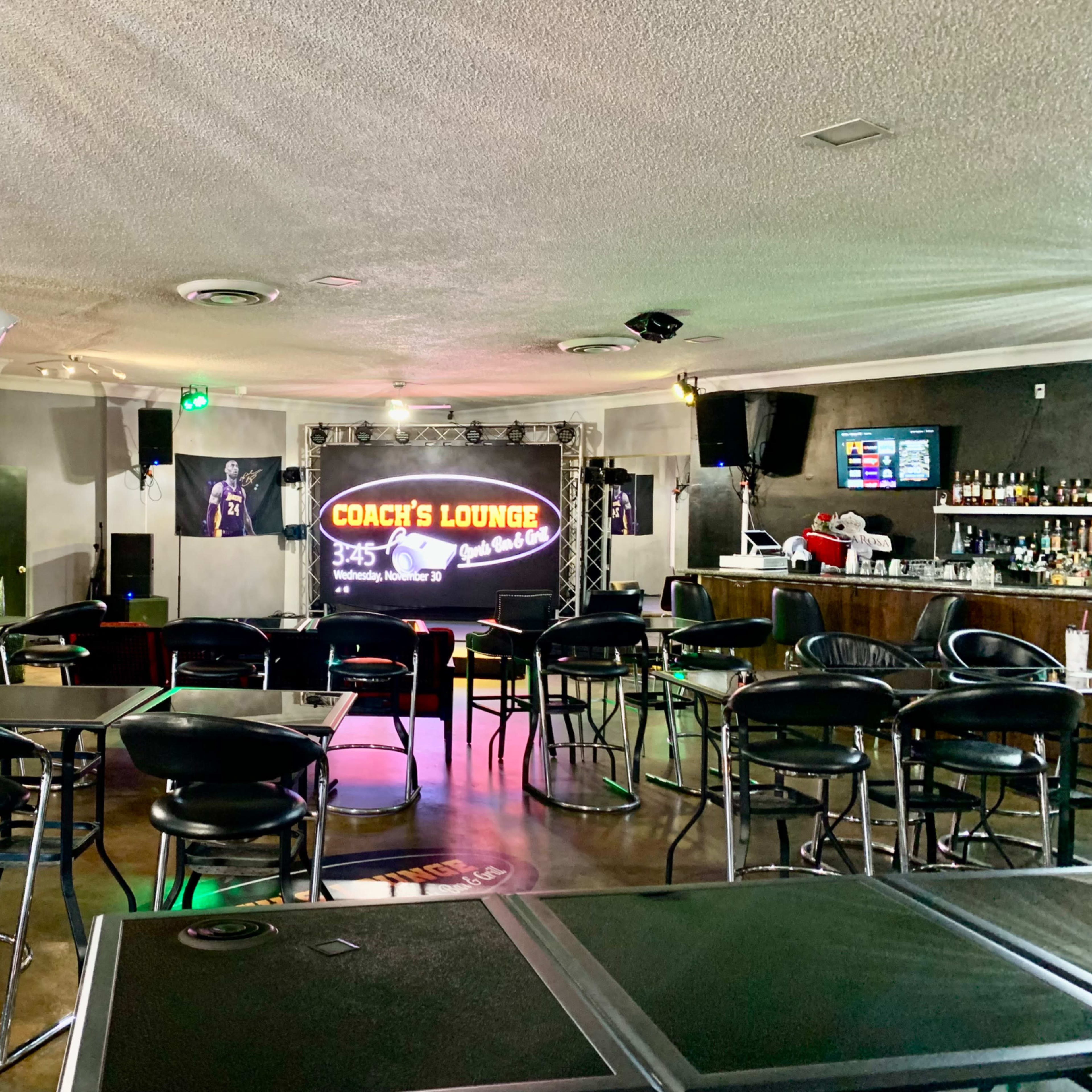 A lounge area with black bar stools and tables, featuring a performance stage with a lighted sign that reads "Coach's Lounge."