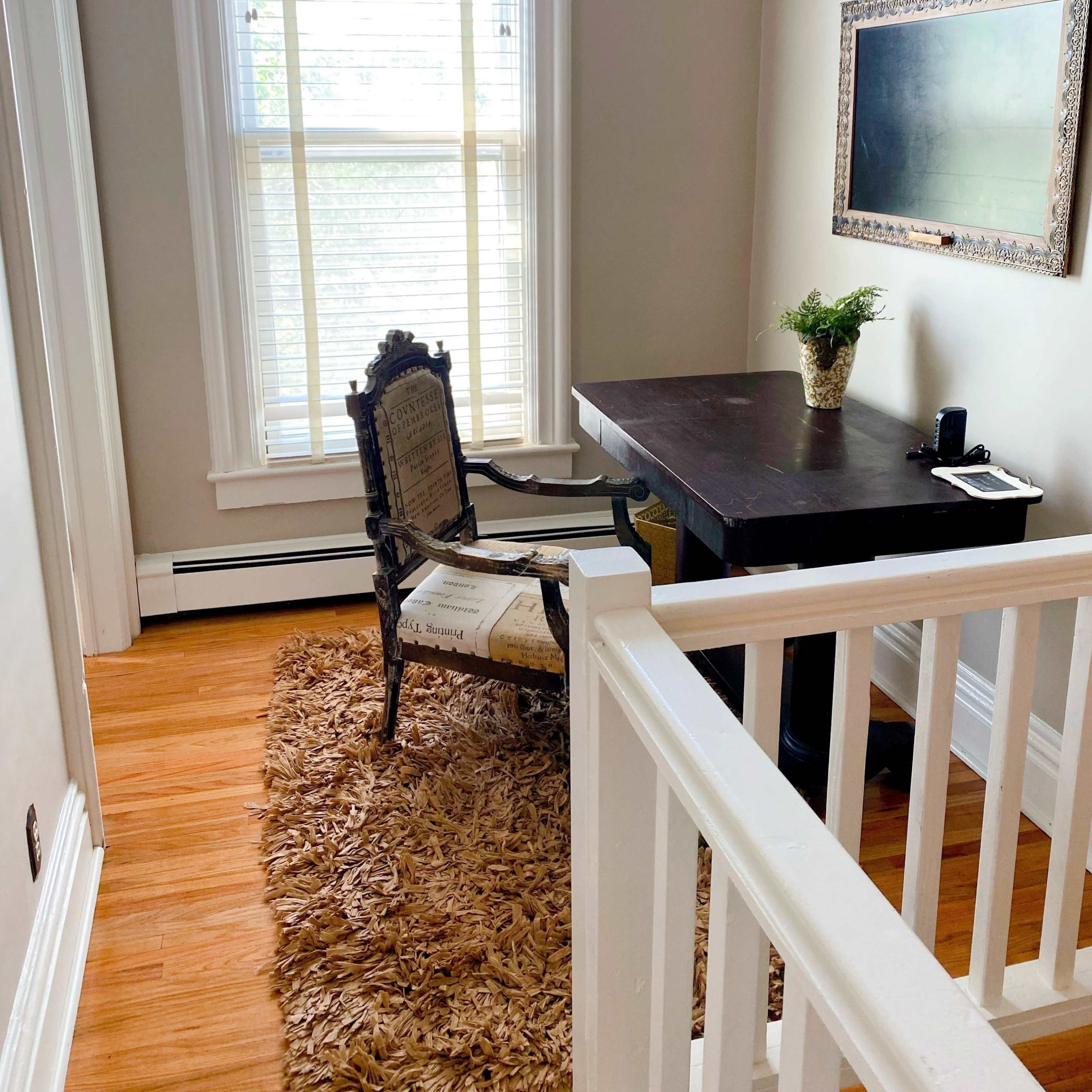 A small, well-lit corner room featuring a dark wooden desk, an ornate chair, and a rug, with a window allowing natural light to enter.
