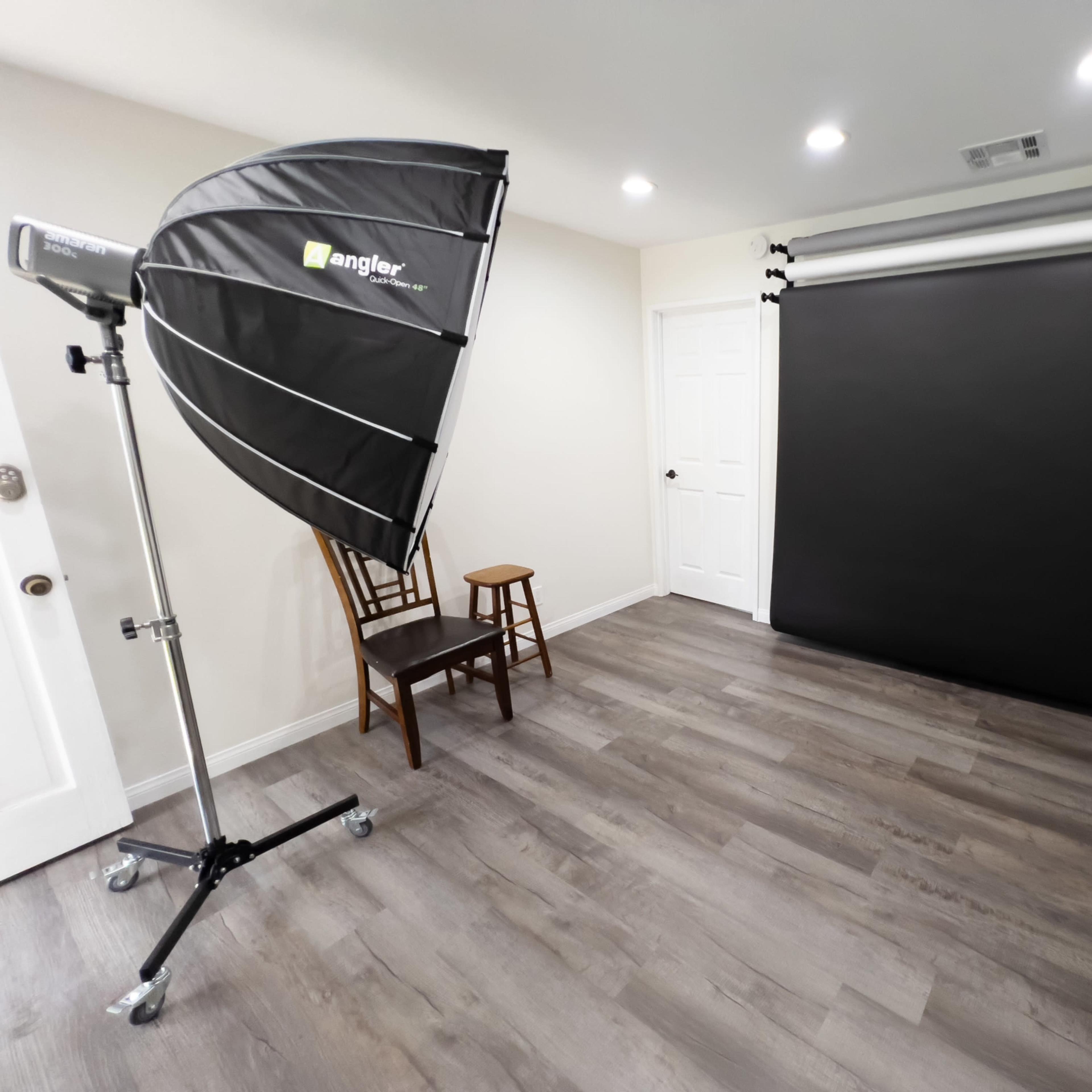 A photography studio with a black backdrop, a light source on a stand, a chair, and wooden flooring.