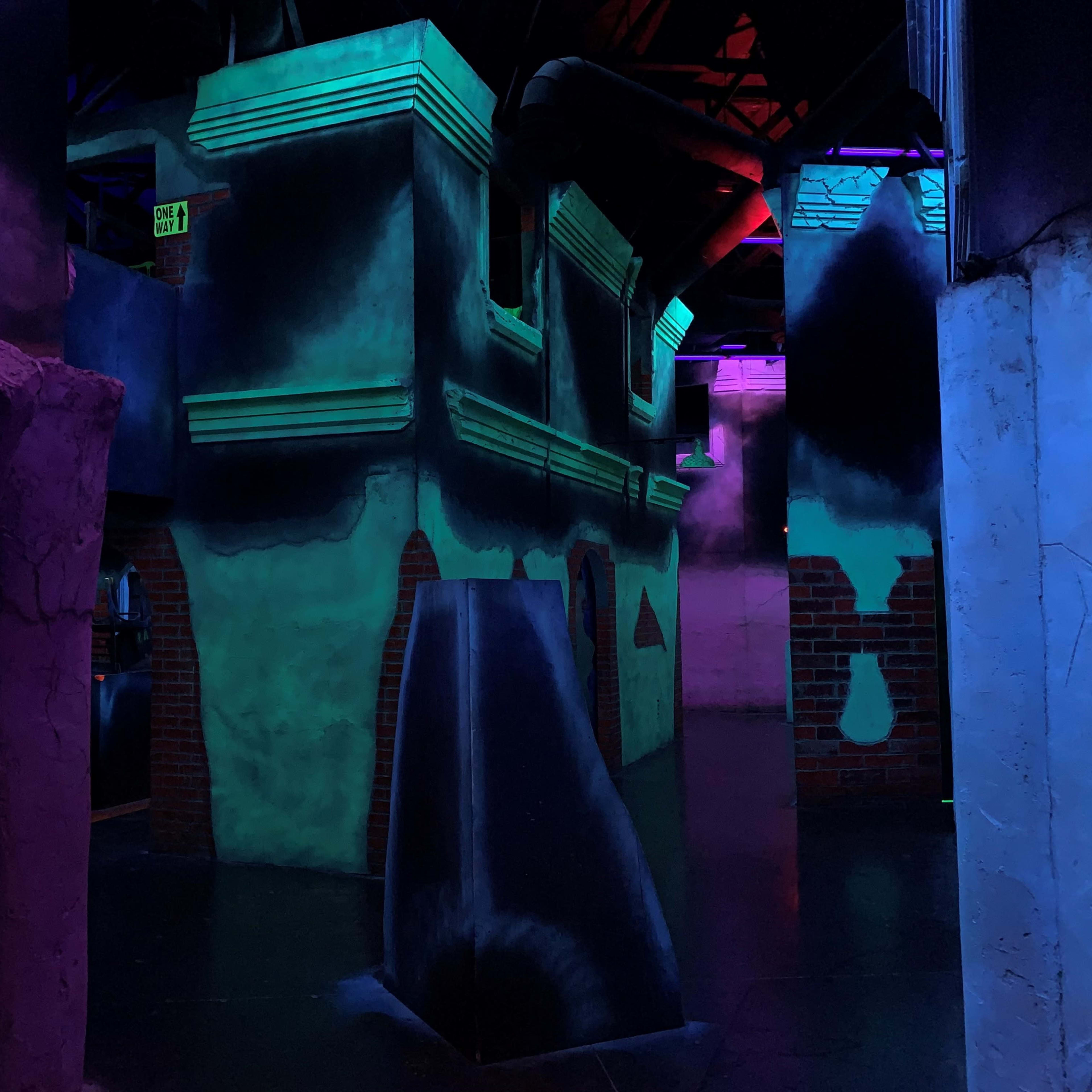 A dimly lit indoor laser tag arena featuring brightly painted walls and various geometric structures.