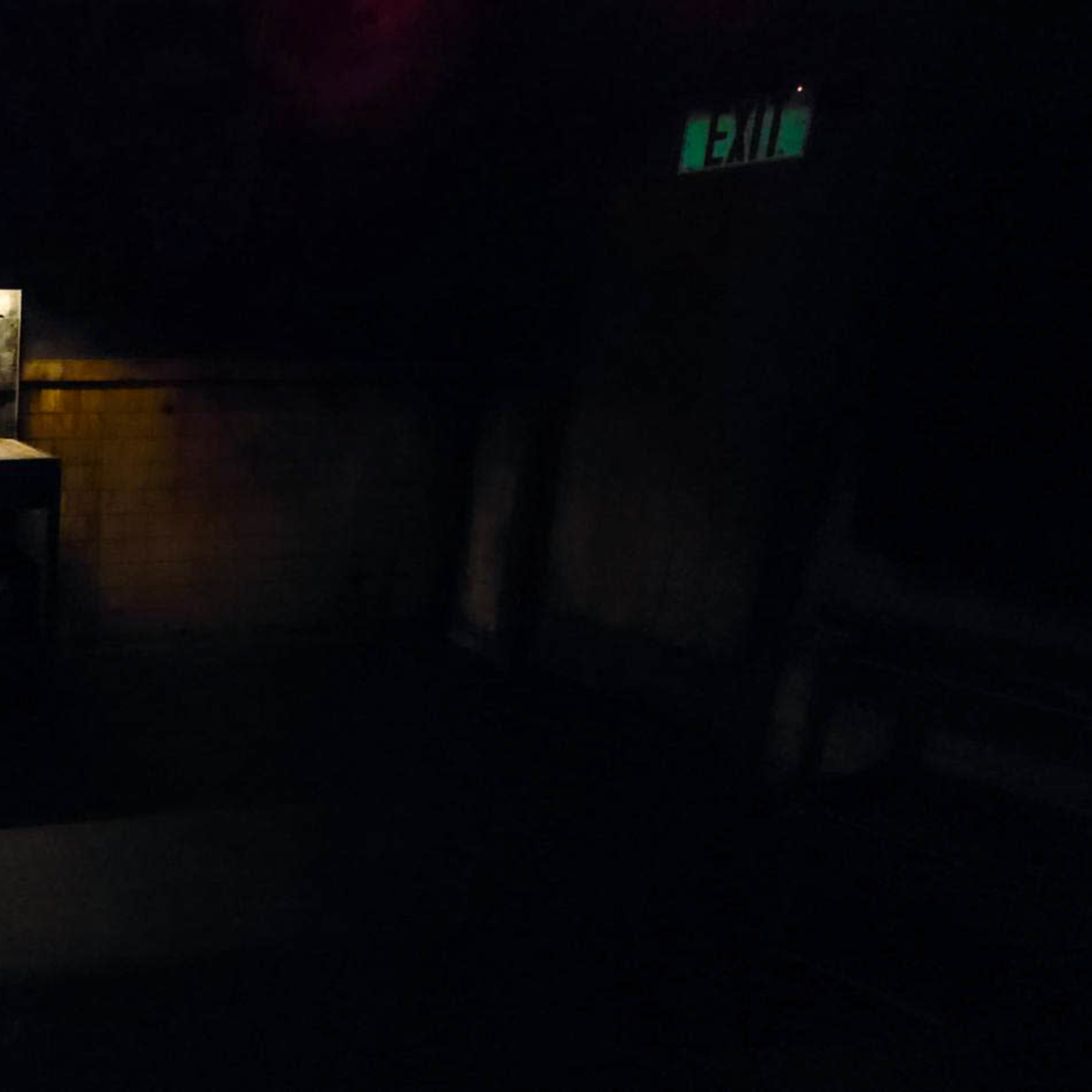 A dimly lit room features a metallic countertop illuminated from above, surrounded by dark walls and a visible exit sign.