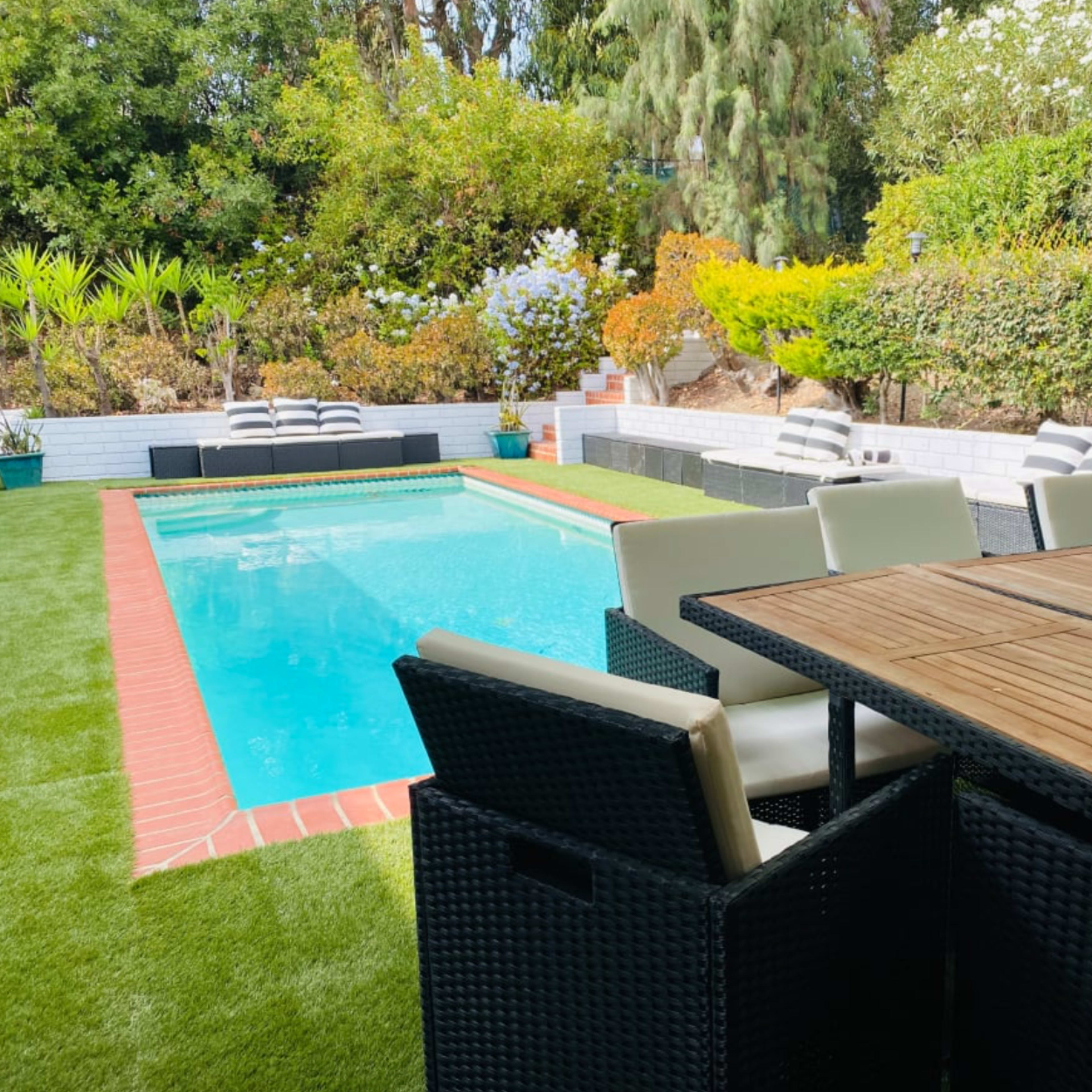 A swimming pool surrounded by a well-maintained garden, with a dining table and chairs positioned nearby.