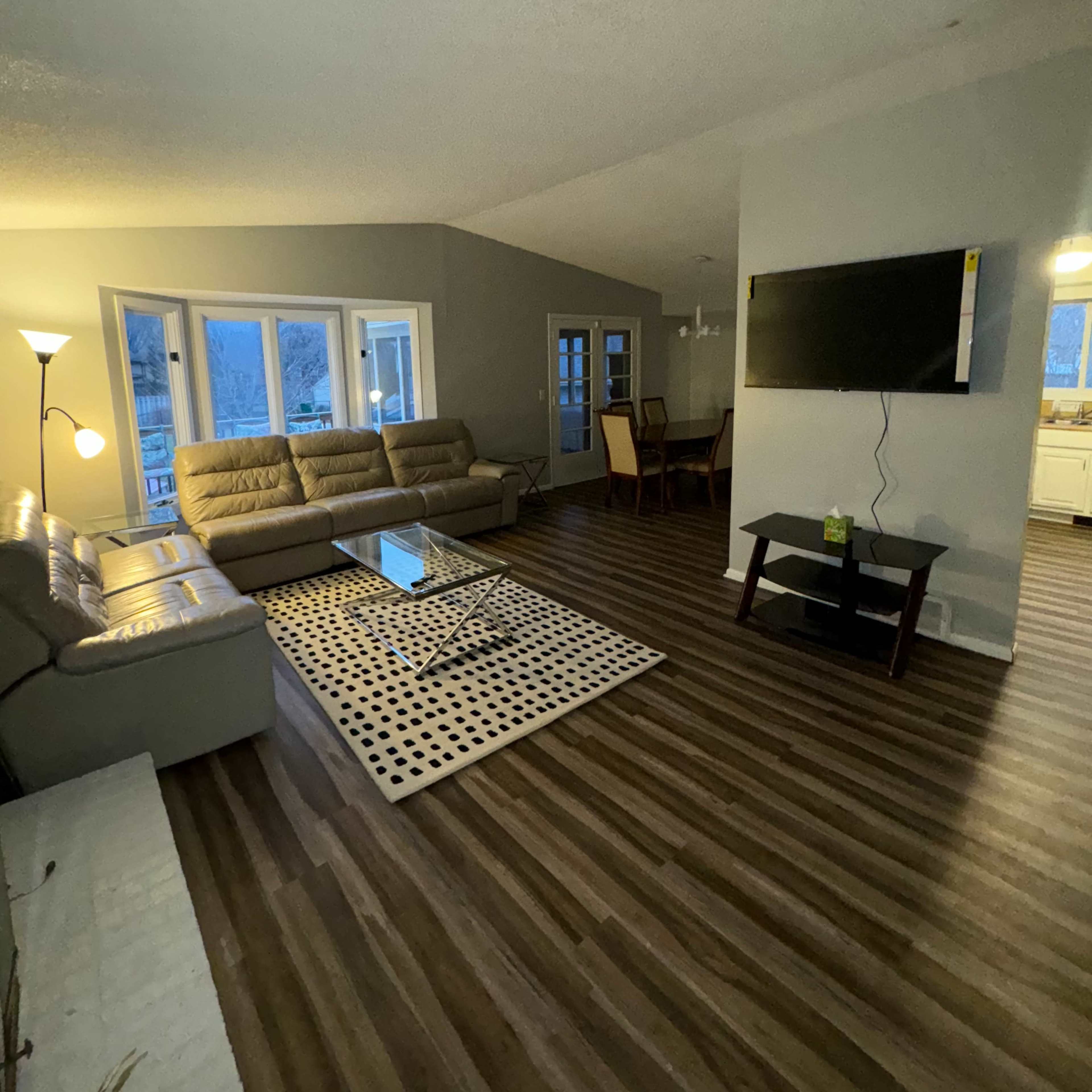 A modern living room with a sectional sofa, a coffee table on a patterned rug, a wall-mounted television, and dining area visible in the background.