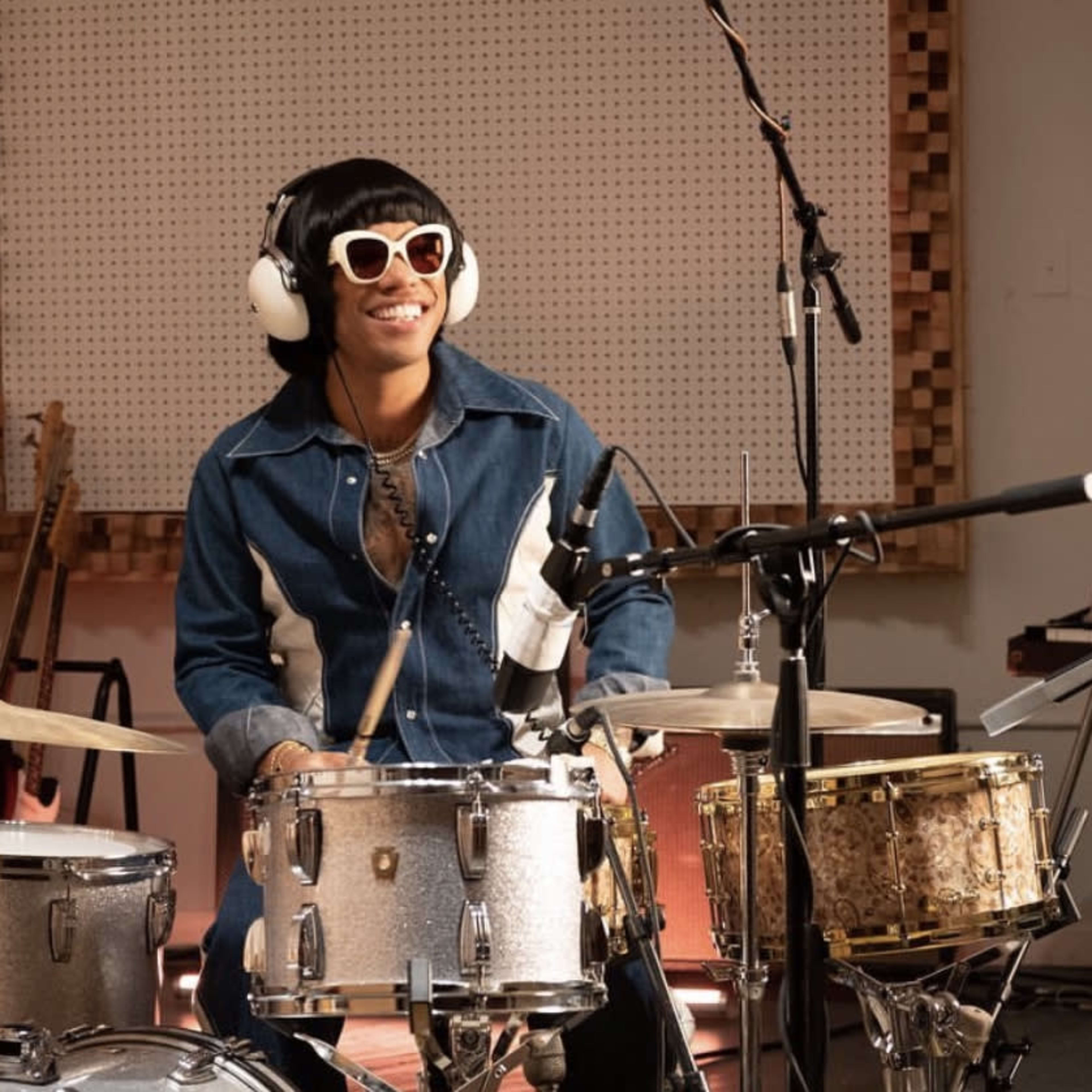 A person wearing sunglasses and headphones plays the drums in a music studio surrounded by instruments.