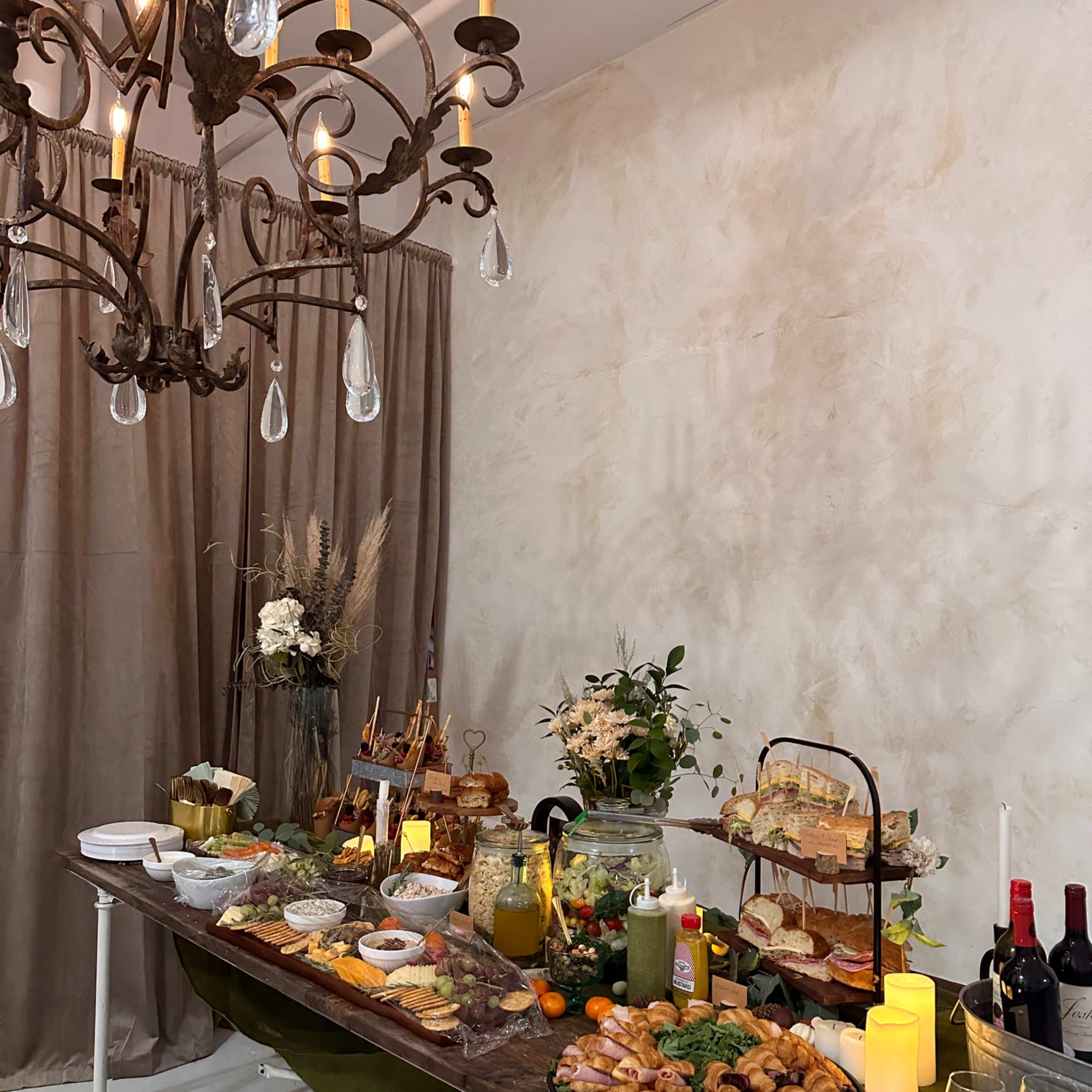A long wooden table is set with various food platters, drinks, and decorative items under a chandelier in a softly lit room.