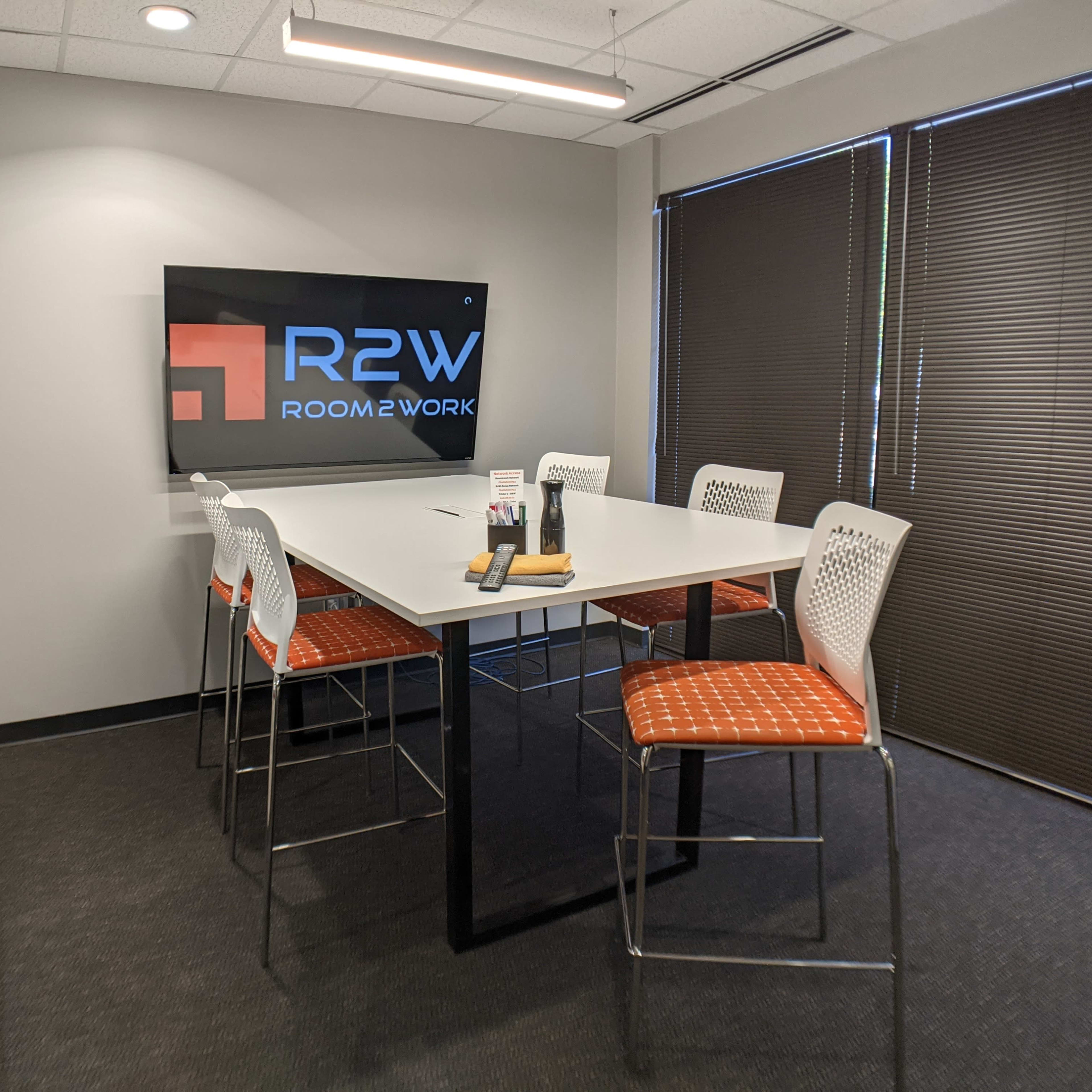 A modern meeting room with a rectangular table surrounded by six high-backed stools and a screen displaying the "R2W ROOM2WORK" logo on the wall.