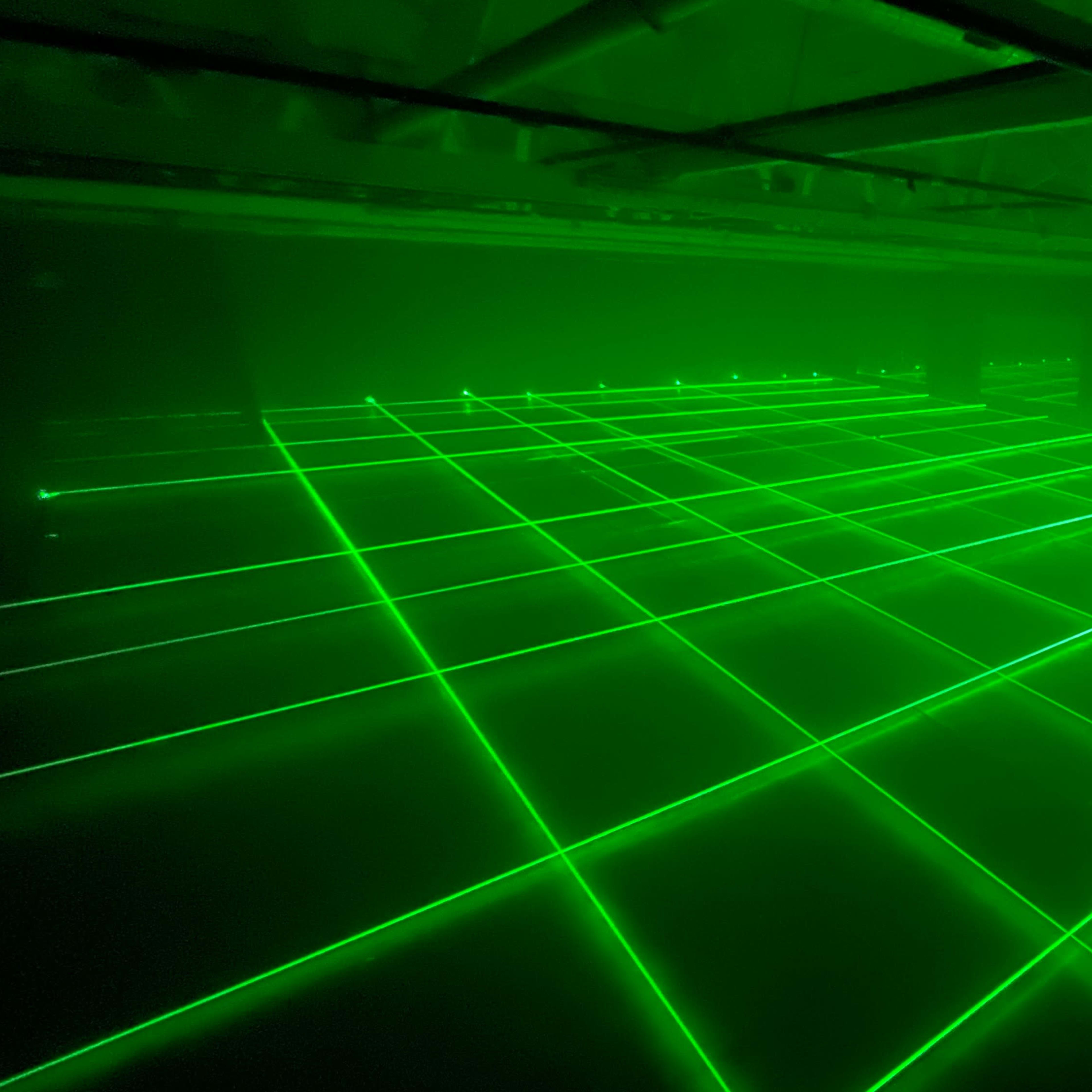 A series of green laser beams are arranged in a grid pattern across a dimly lit space.