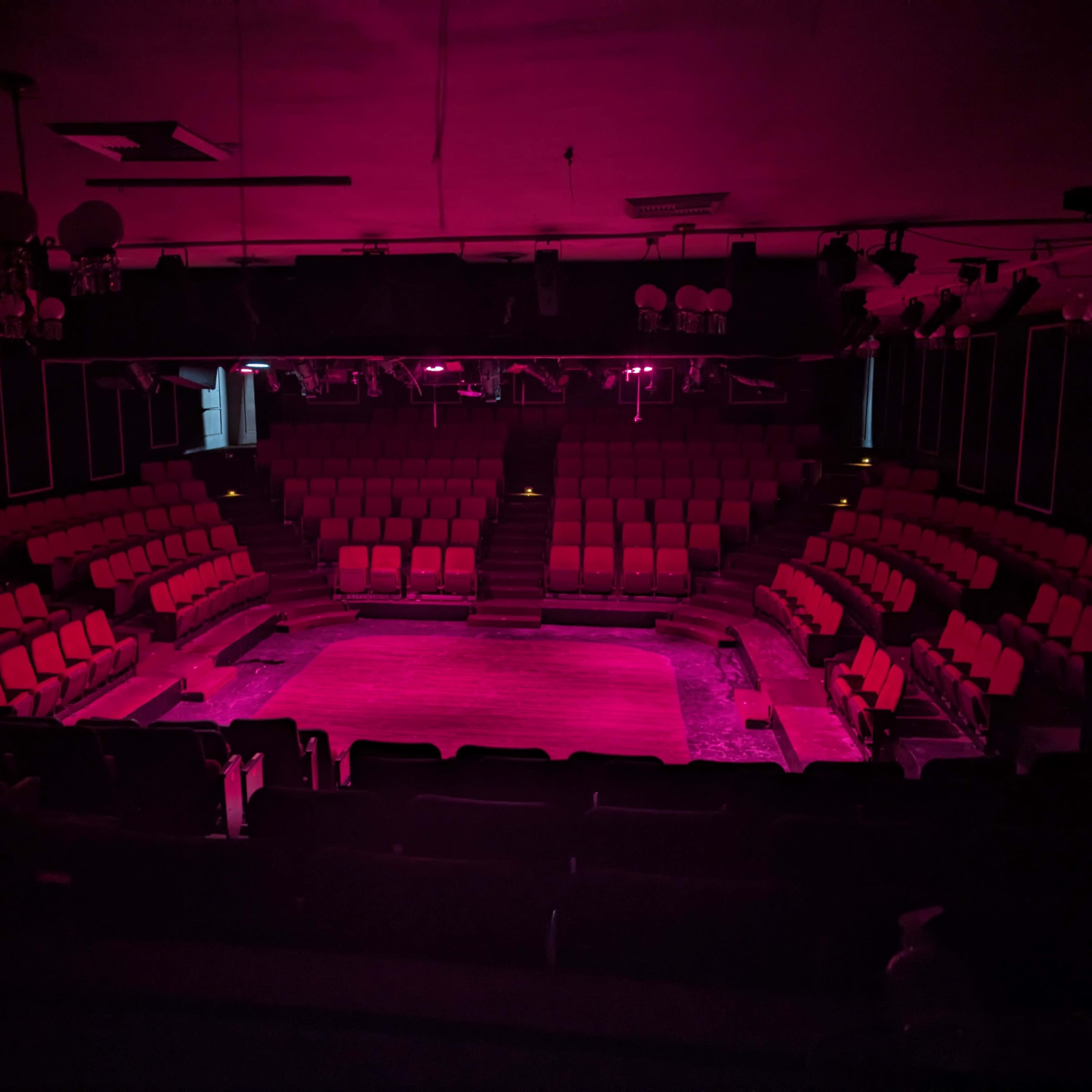 A dimly lit theater with red lighting and empty seats arranged around a central stage.