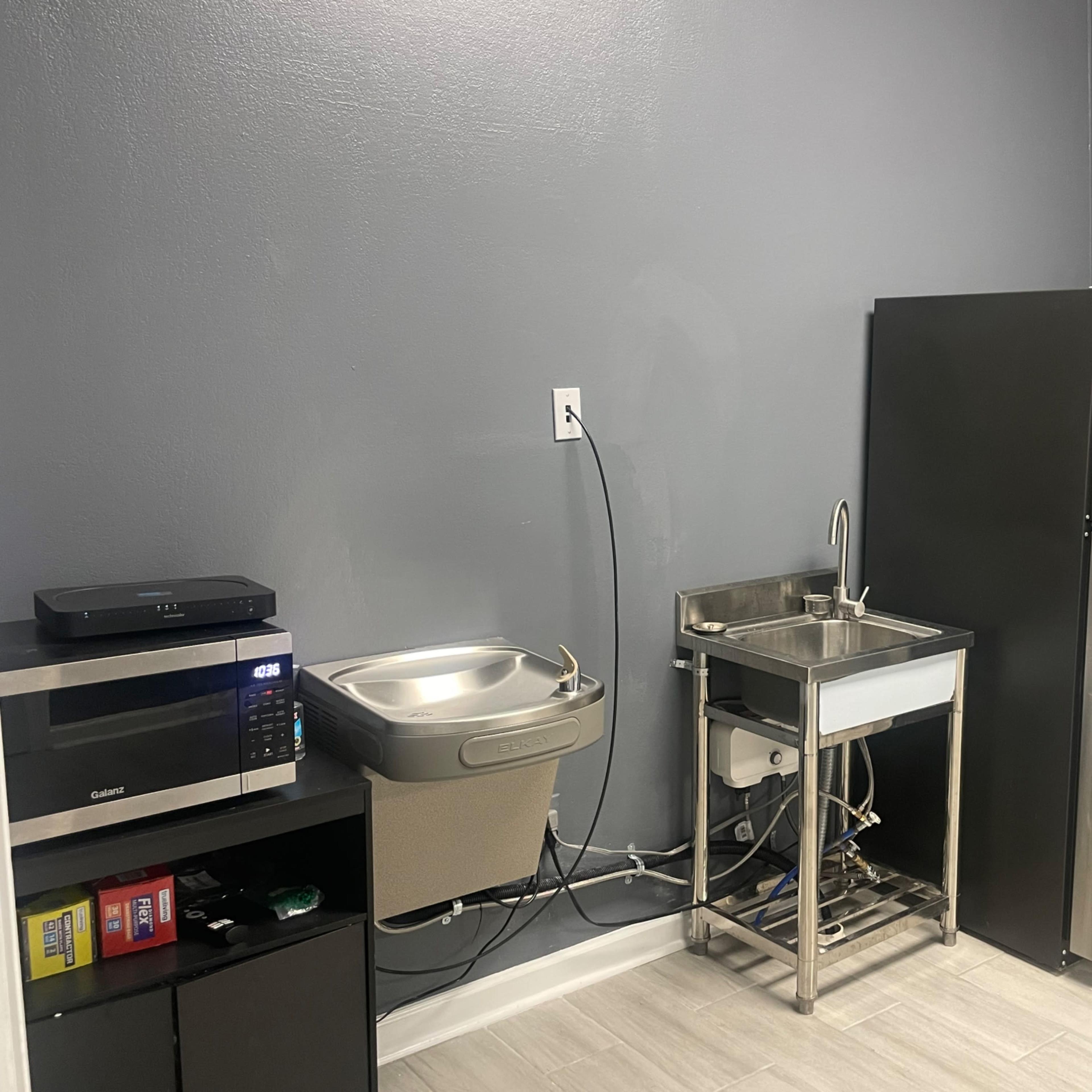 A kitchen area with a stainless steel sink, a refrigerator, a microwave, and a waste bin.