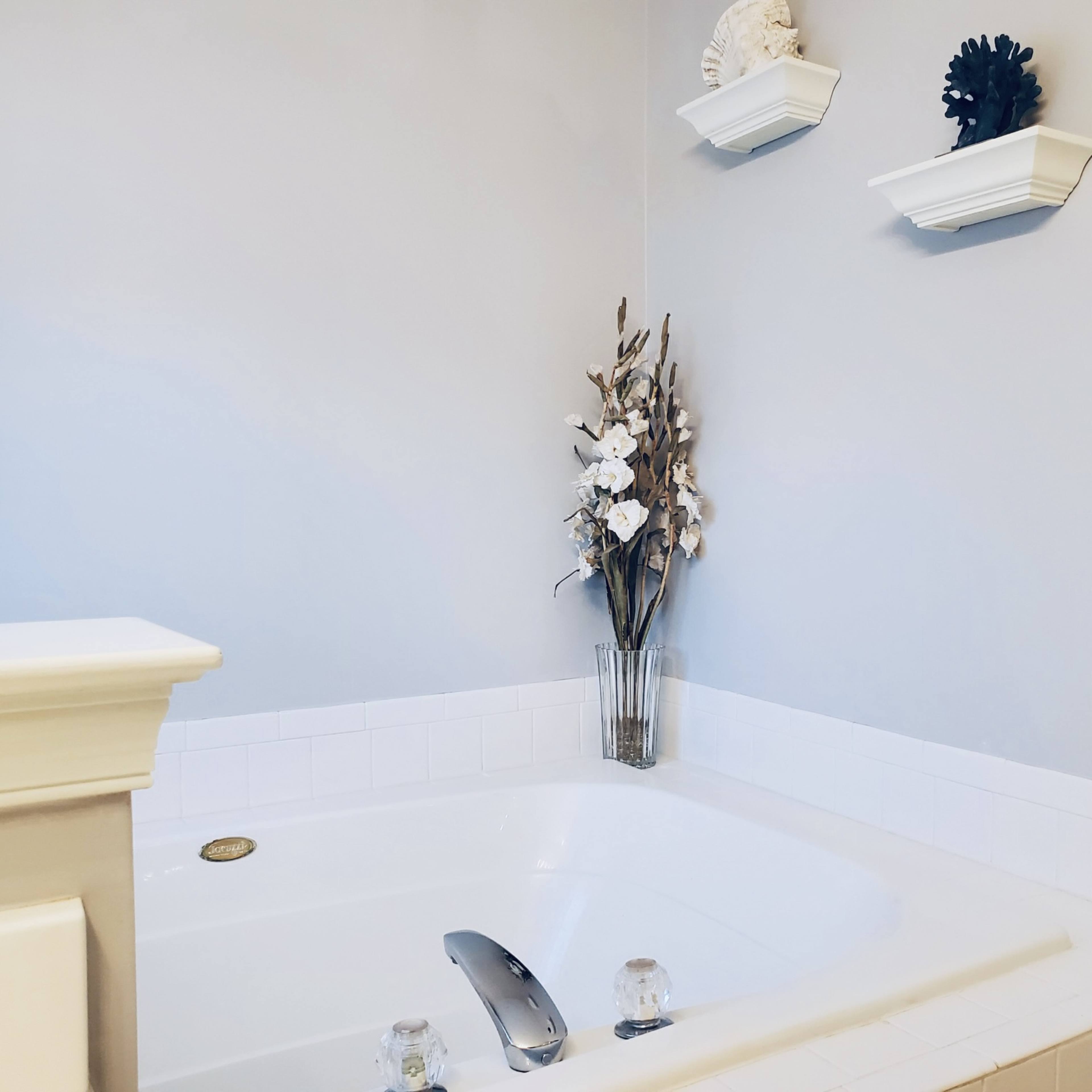 A corner of a bathroom featuring a white jetted tub accompanied by a clear vase with decorative flowers and two wall-mounted shelves.