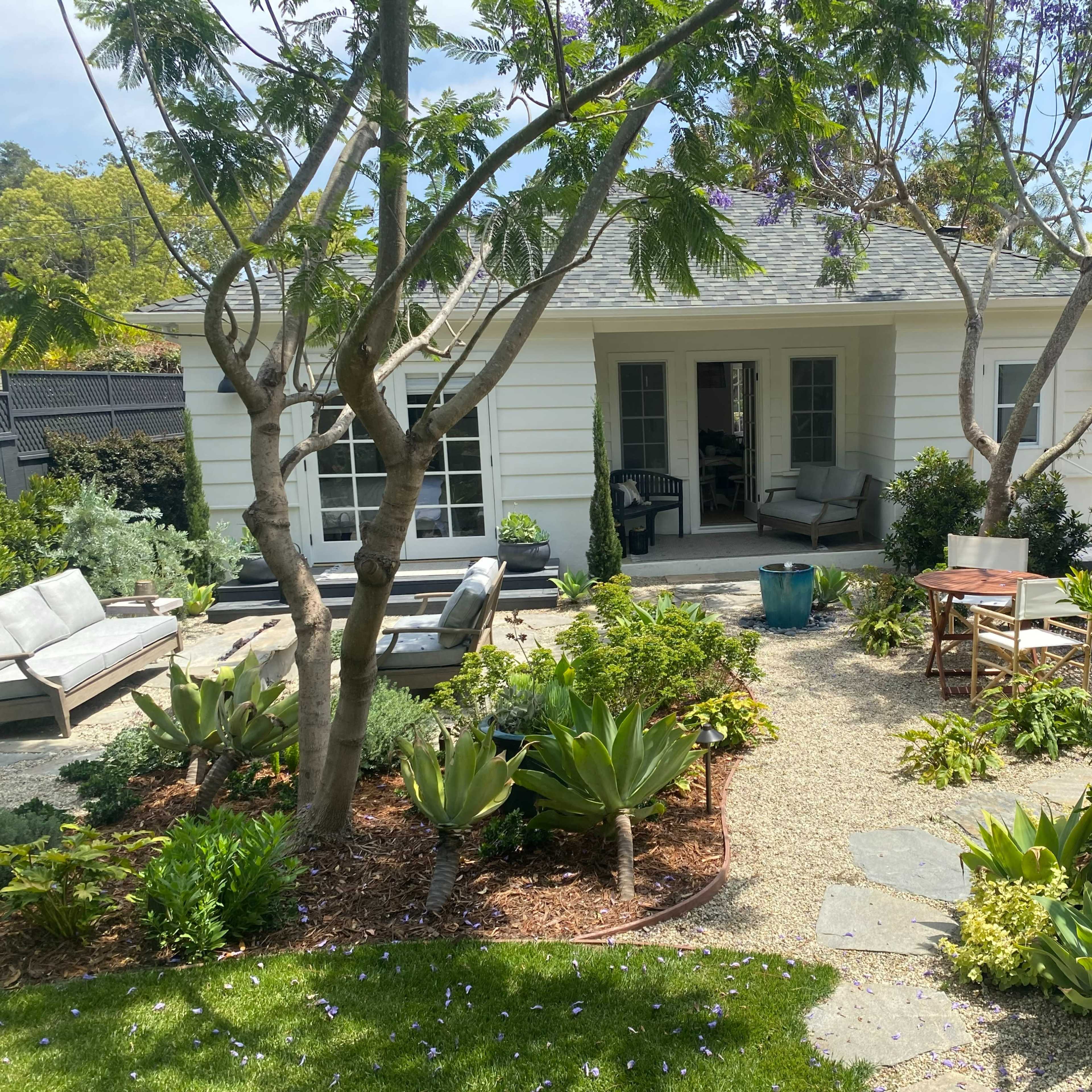 A backyard garden features a seating area with sofas, a small table, and a staircase leading to a second level, surrounded by various plants and trees.