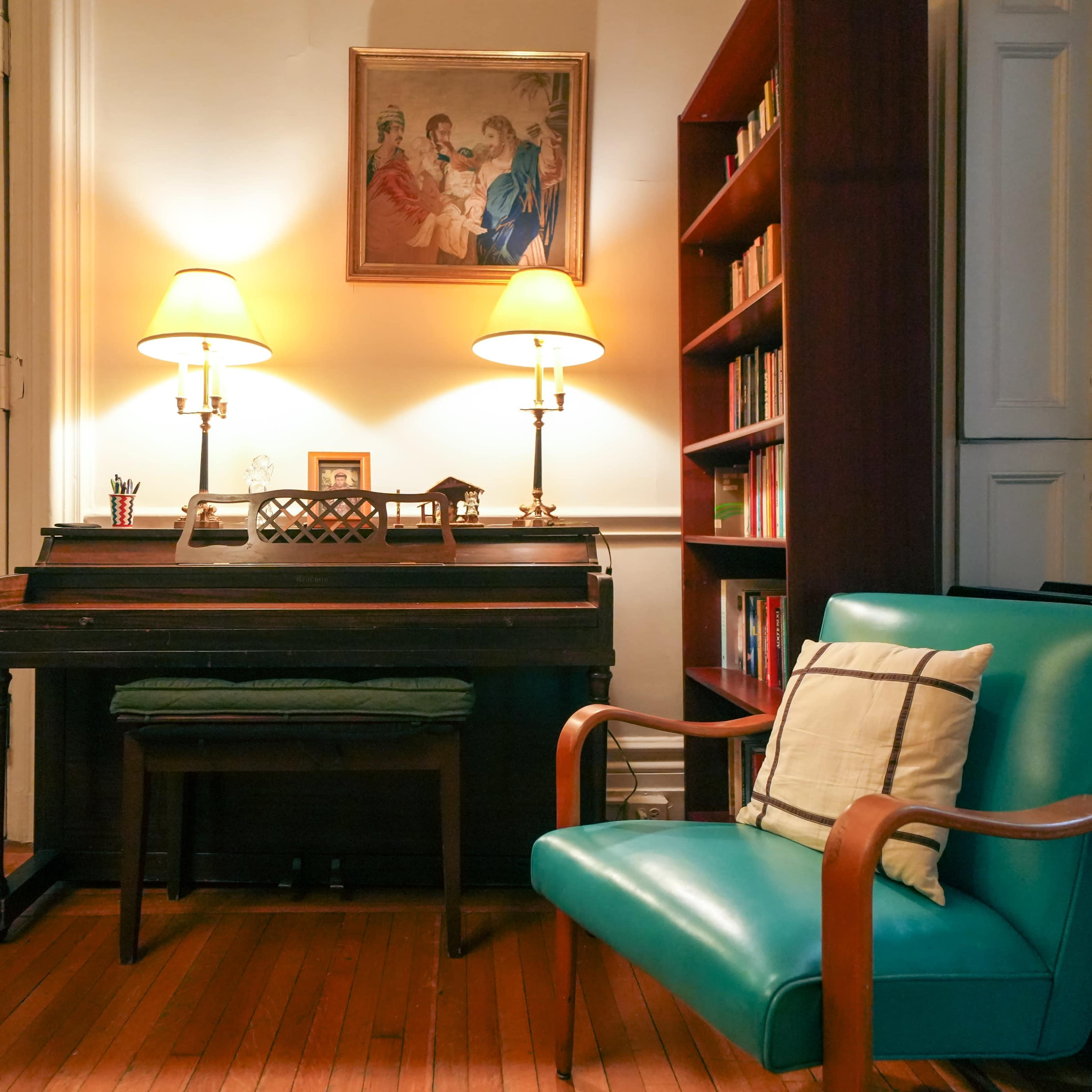 A cozy corner of a room featuring a wooden desk with a piano, two lamps illuminating the space, and a teal chair beside a bookshelf filled with books.