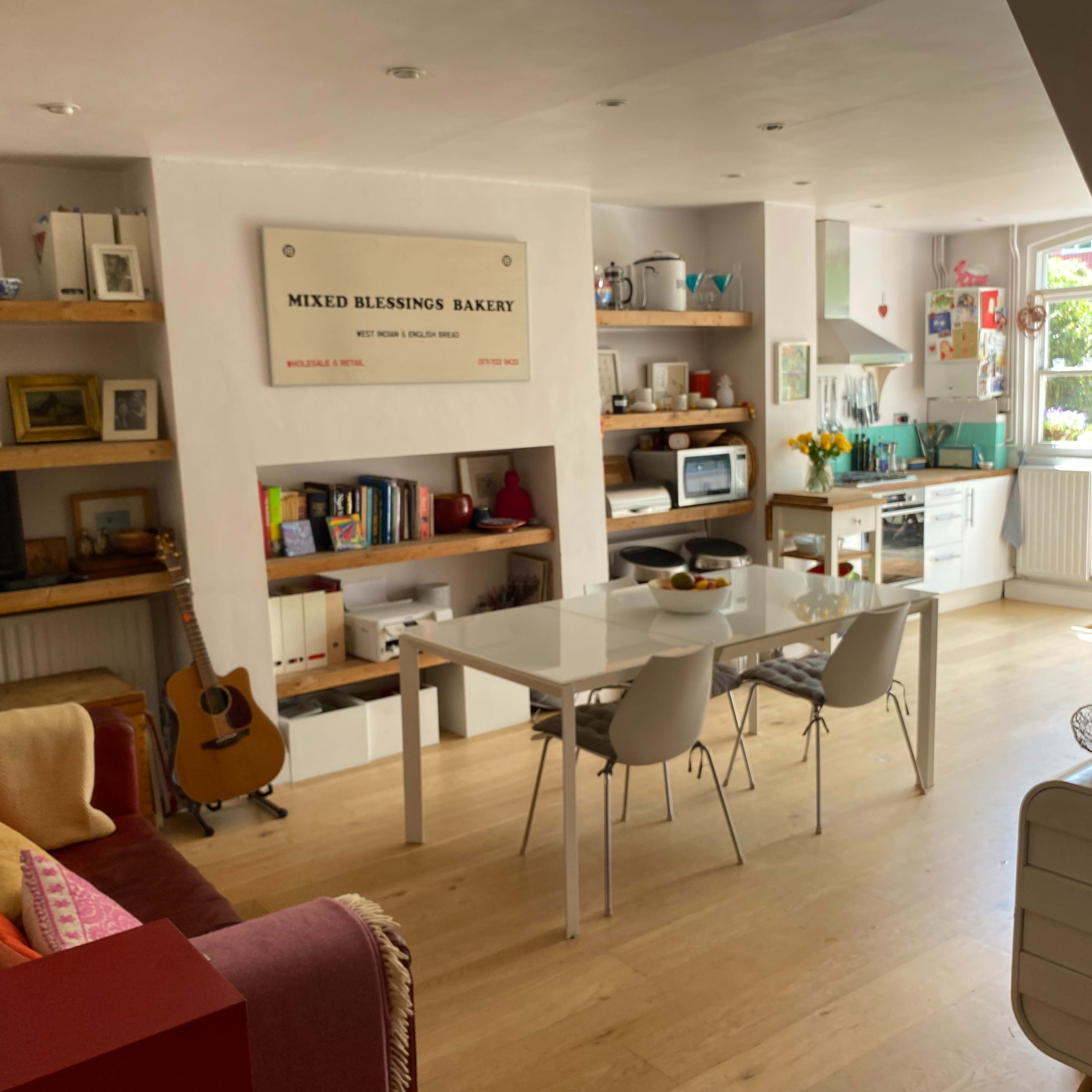 A bright and modern kitchen and dining area featuring a white table with chairs, wooden shelves with decorative items, and a guitar leaning against a wall.