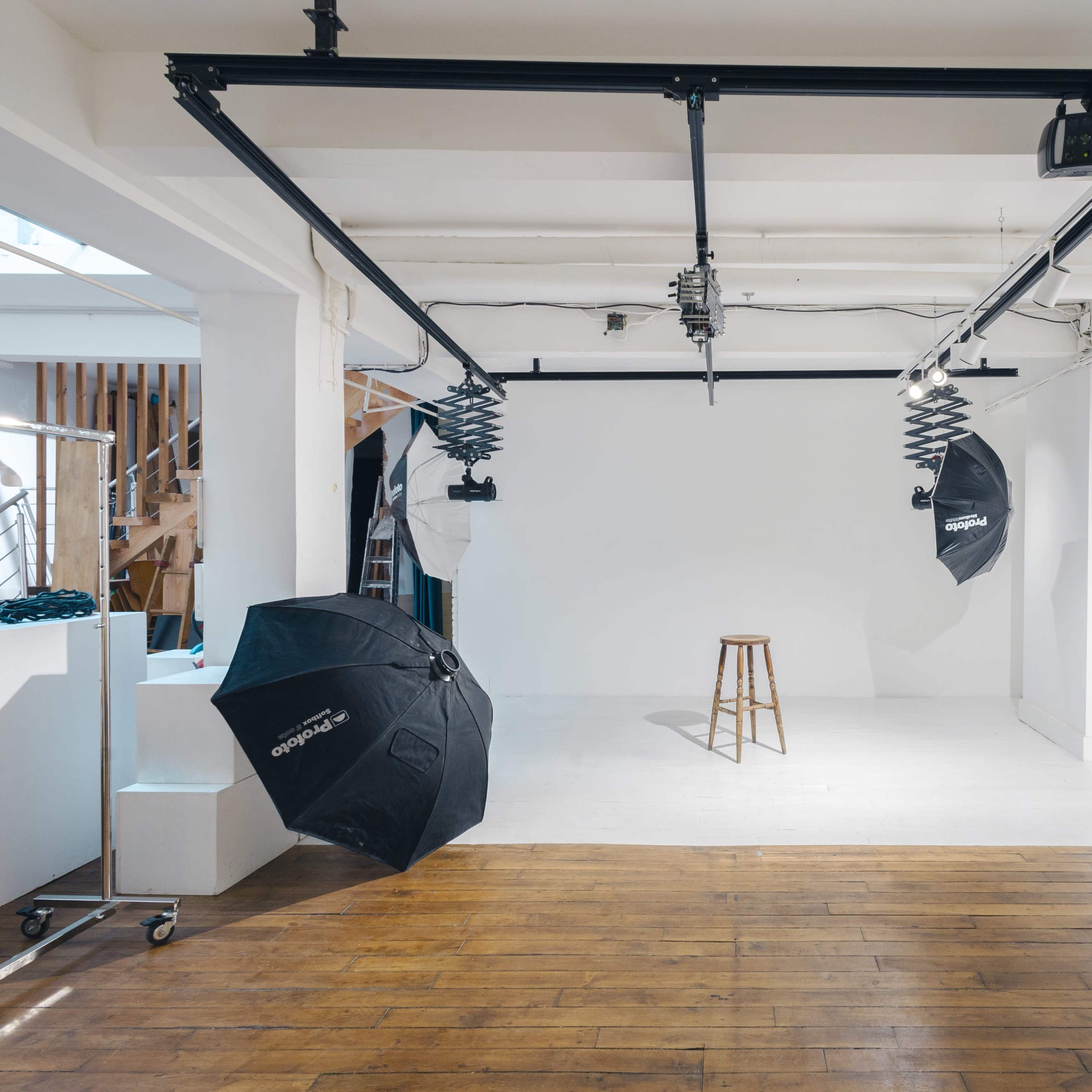 A photography studio features a white backdrop, a wooden stool, and several light umbrellas positioned around the space.