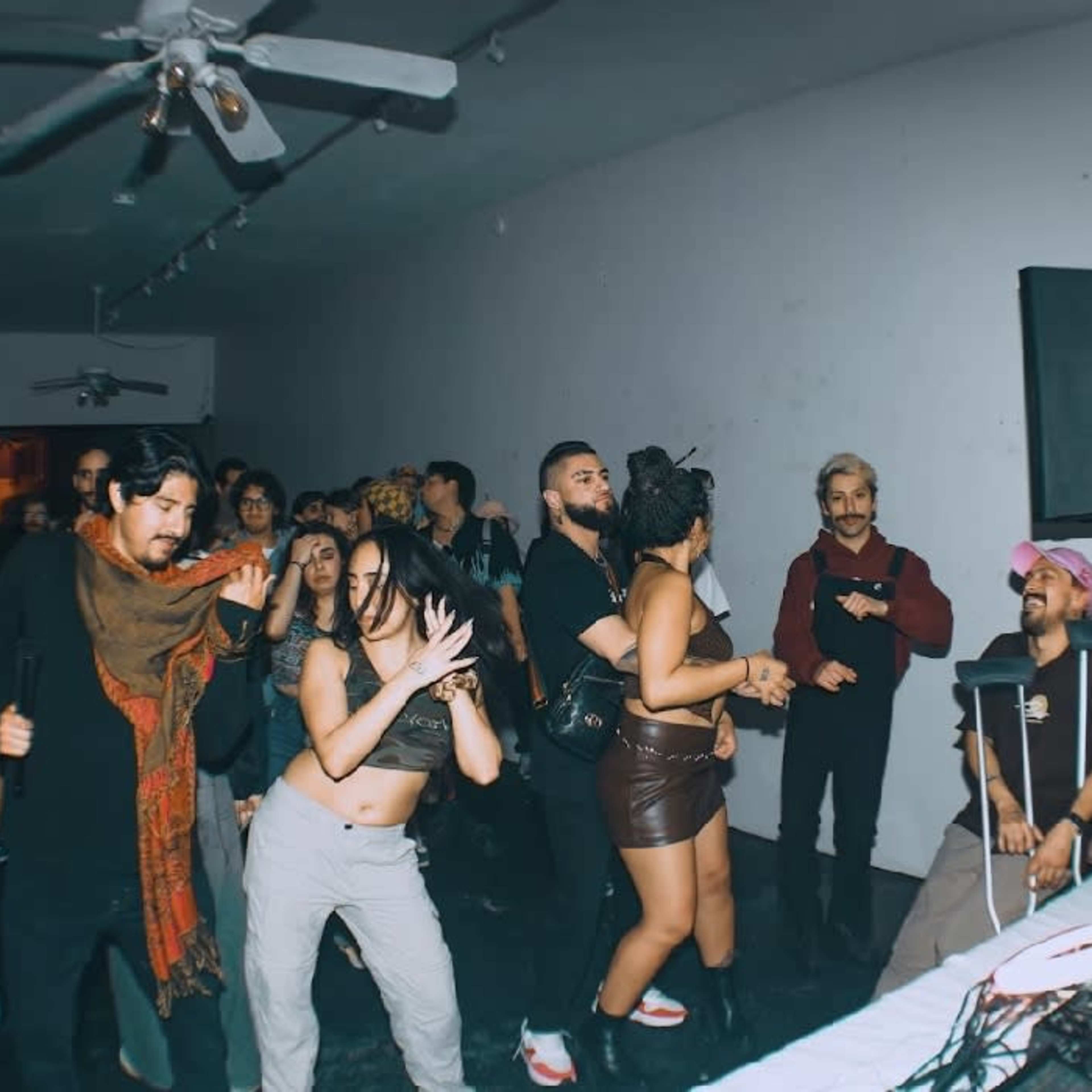 A group of people dances in a dimly lit space with a DJ setup and sound equipment in the foreground.