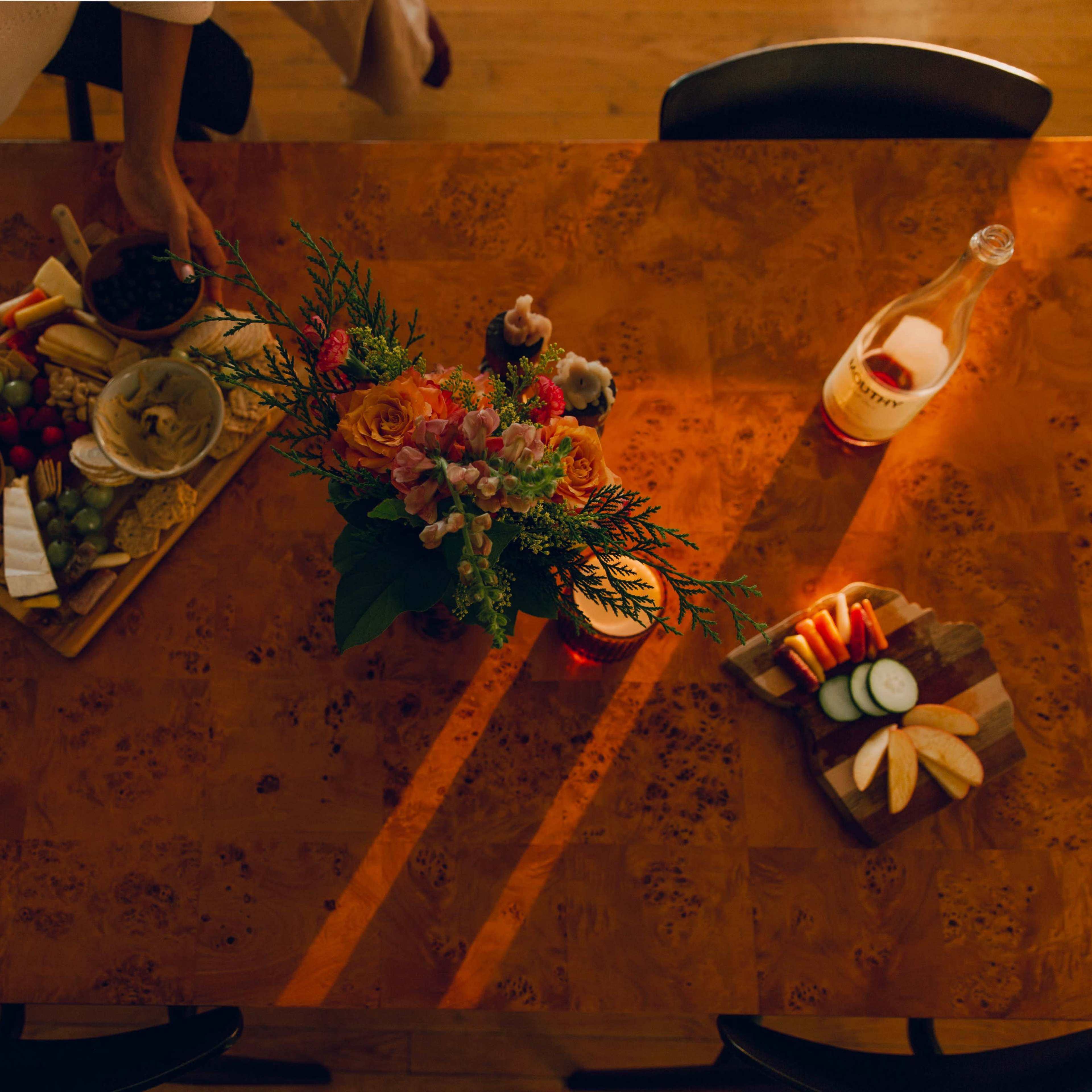 A person arranges a platter of cheese, fruit, and dip beside a vase of flowers on a wooden dining table.