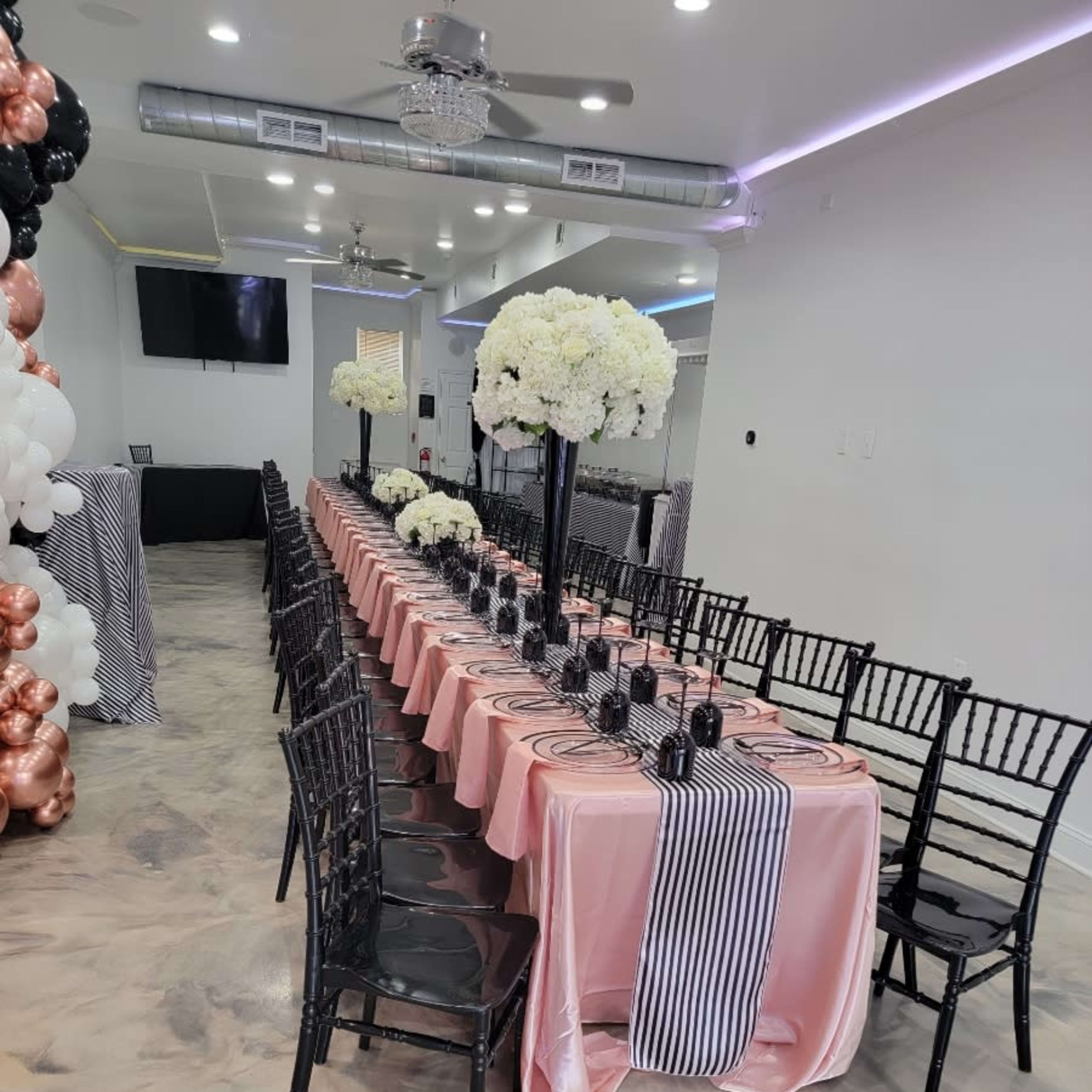 A long dining table is elegantly set with pink tablecloths, black plates, and large white floral centerpieces in a decorated room.
