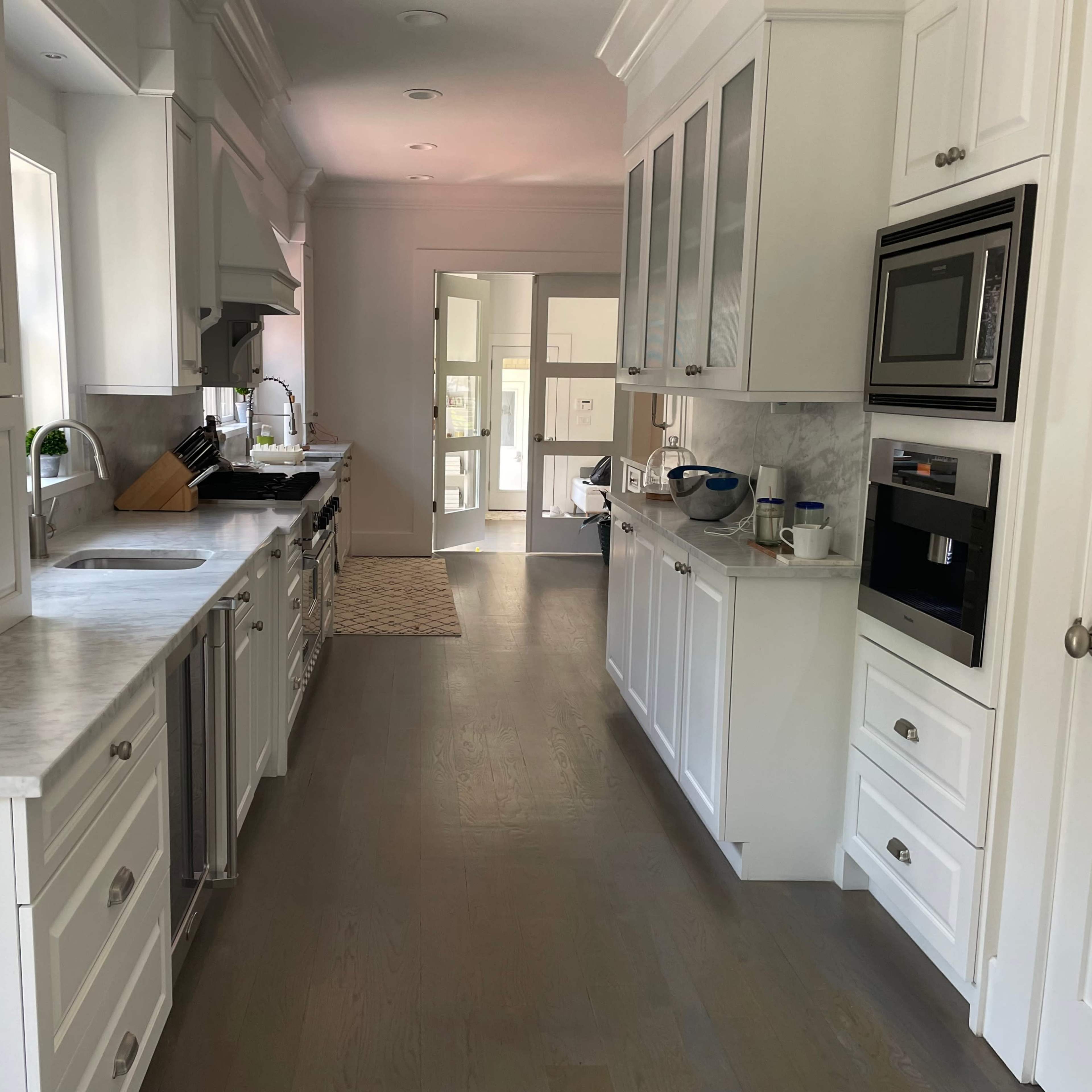 A modern kitchen with white cabinetry, marble countertops, and stainless steel appliances, extending into a spacious hallway.