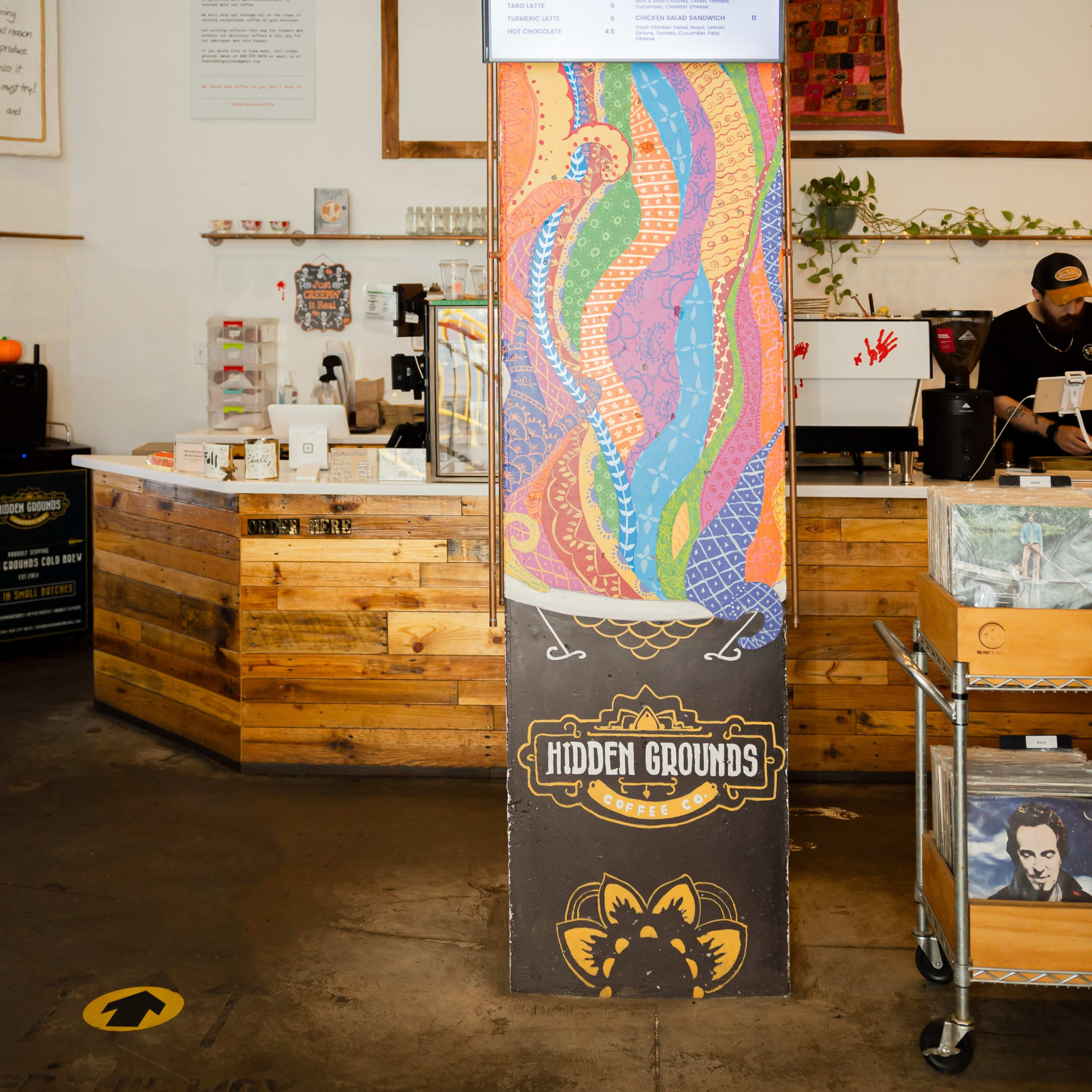 The interior of a coffee shop featuring a colorful mural on a central pillar, a wooden counter, and a menu displayed above.