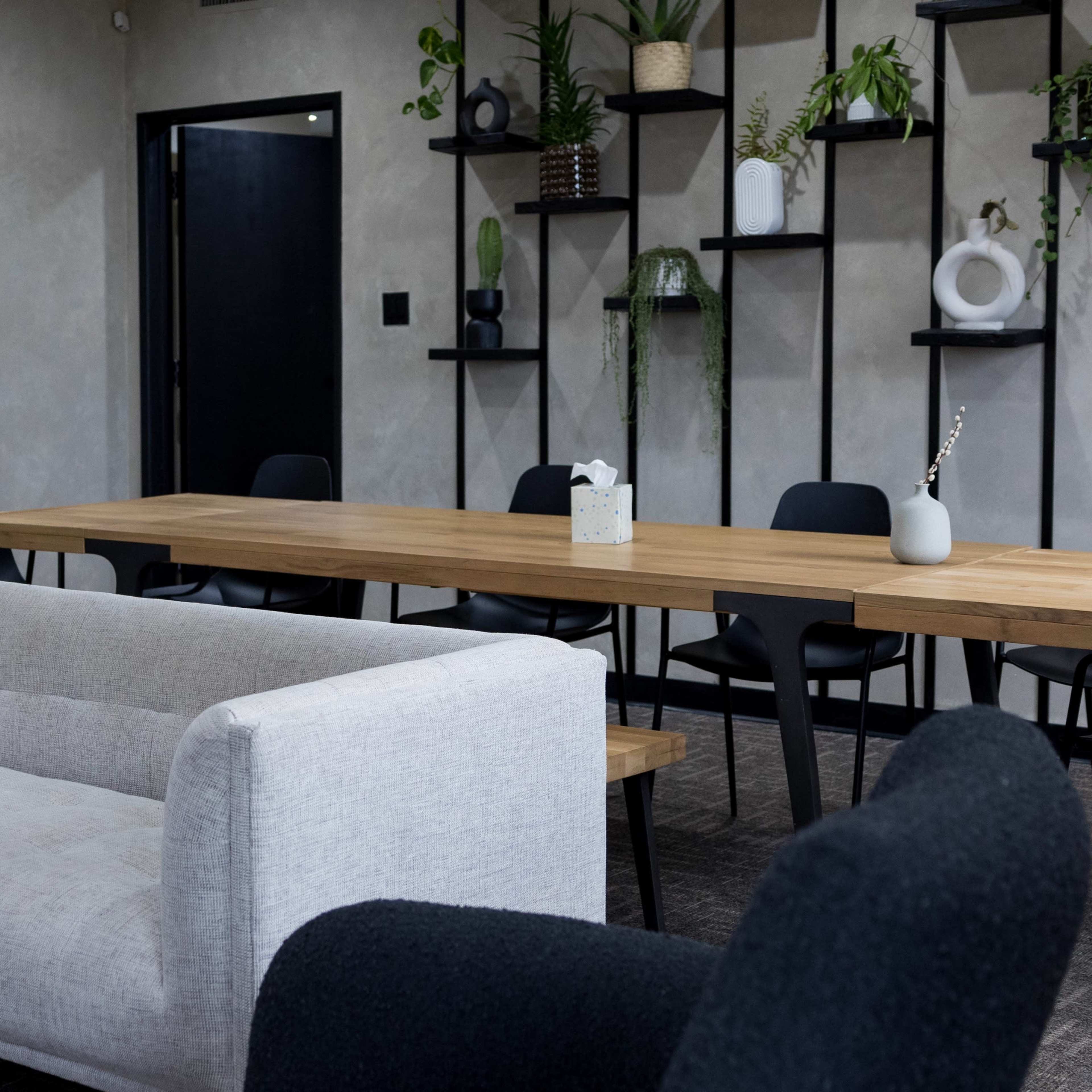 A modern meeting room features a light gray couch and a long wooden table surrounded by black chairs, with plants displayed on shelves along the wall.