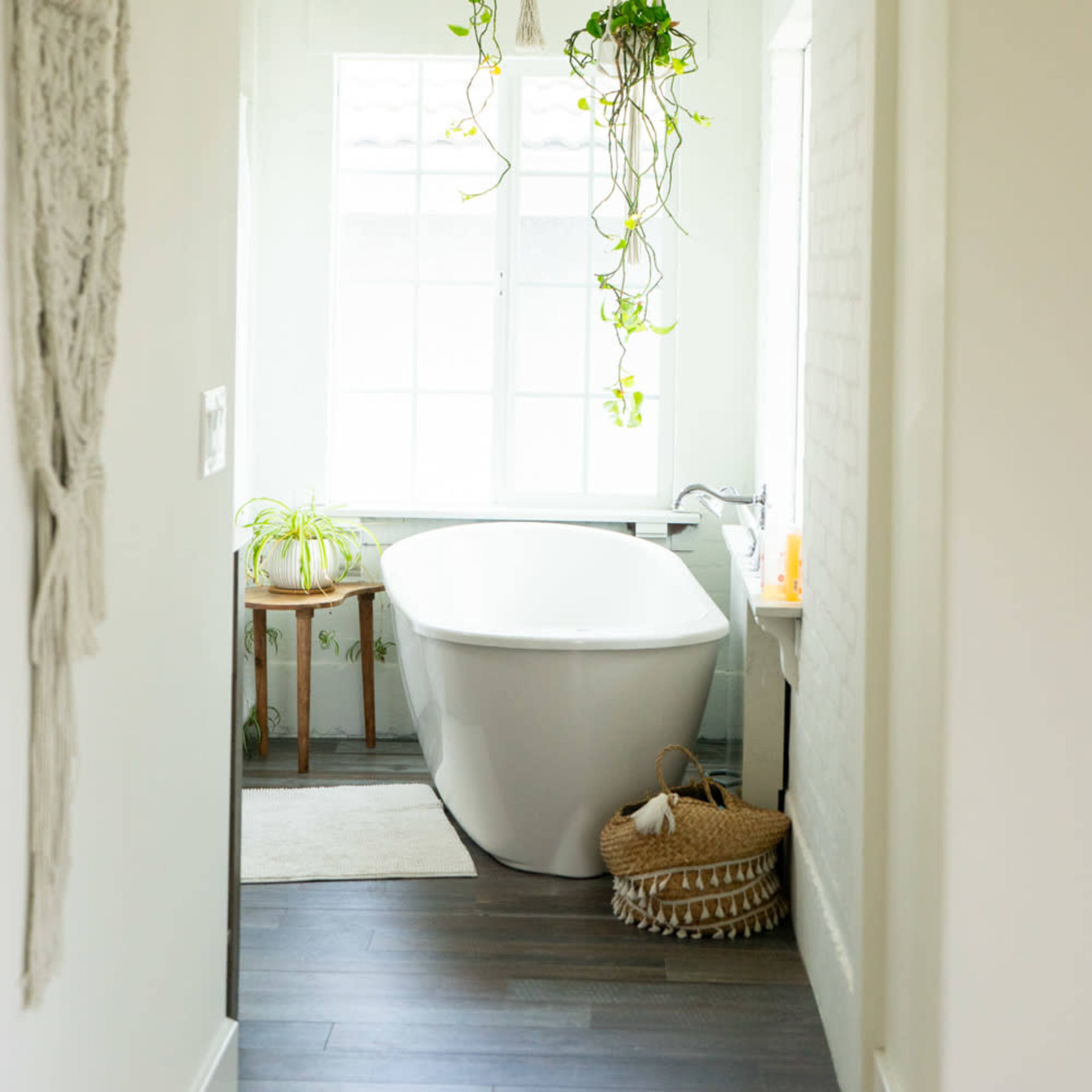 A modern bathroom features a freestanding bathtub with hanging plants above and a window offering natural light.