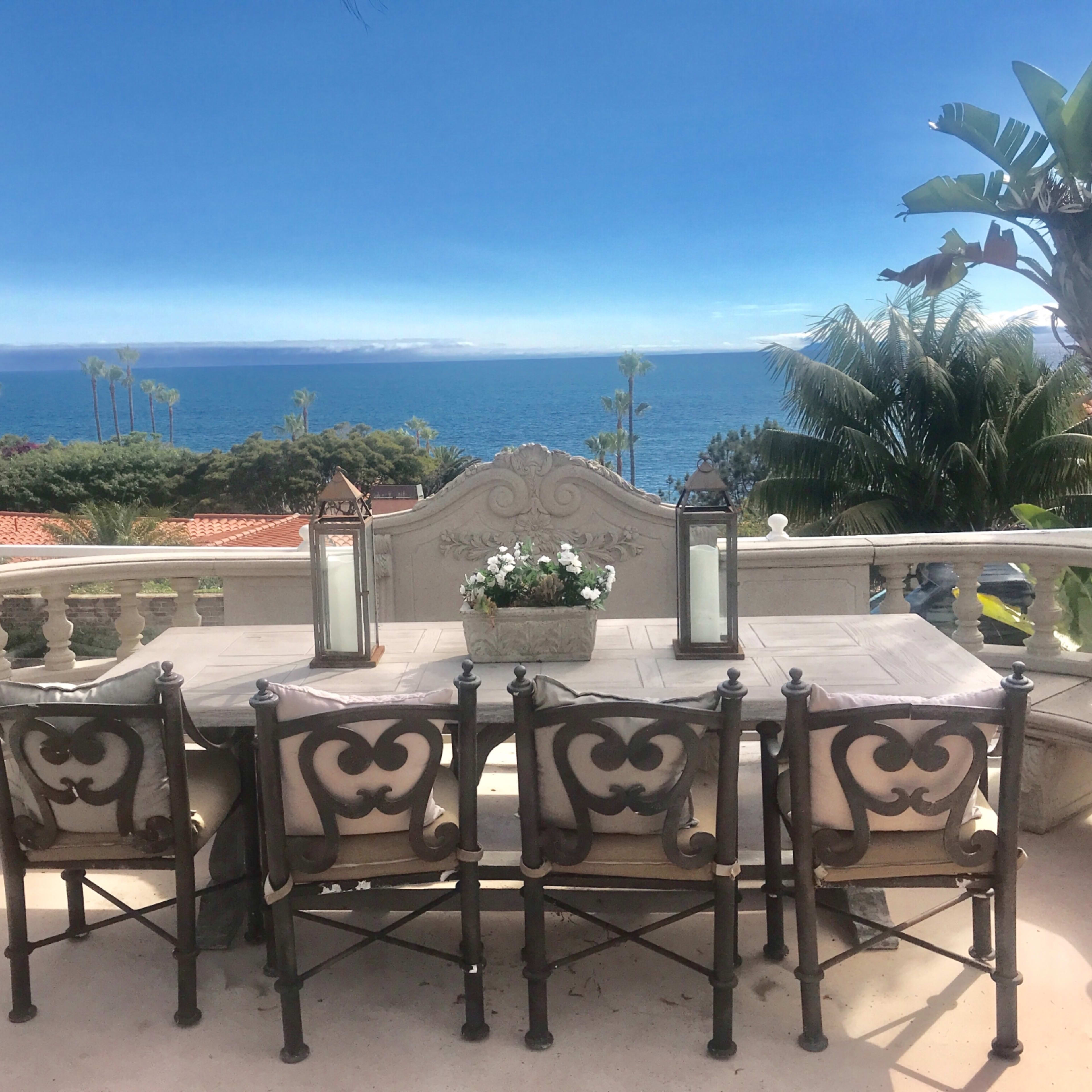 A dining table with six chairs is set on a terrace overlooking the ocean, framed by palm trees and plants.
