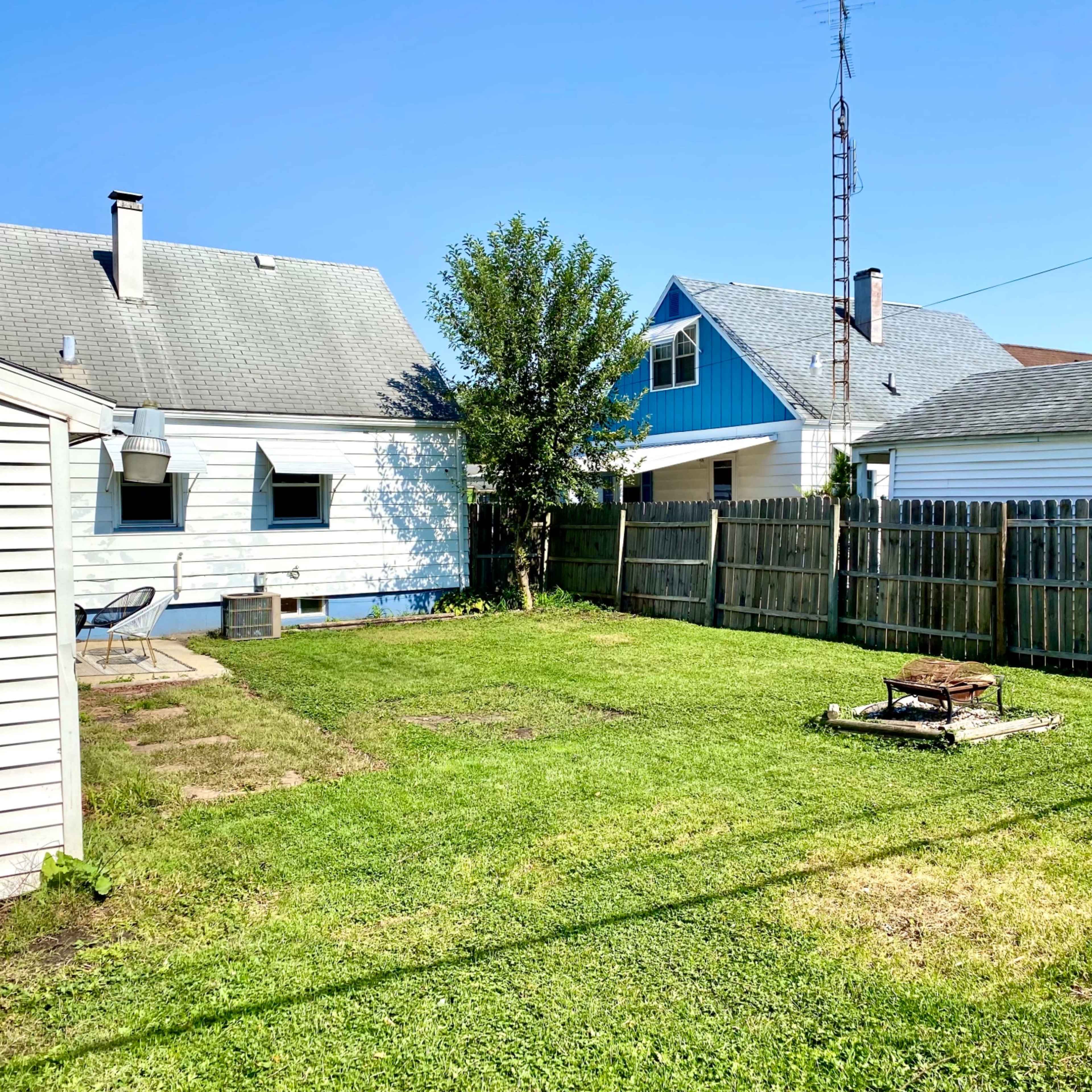 A grassy backyard enclosed by a wooden fence, with two houses in the background and a fire pit in the center.