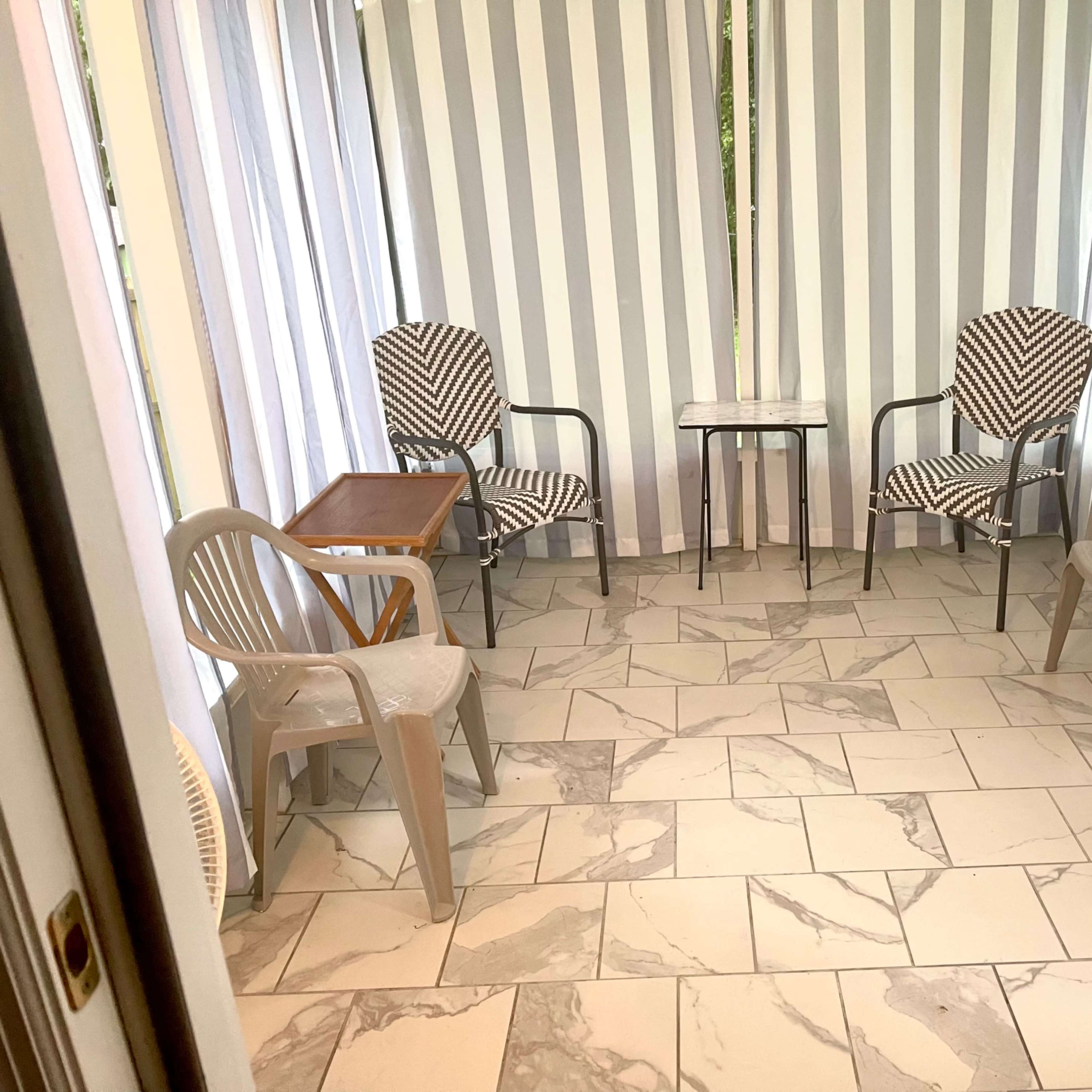 A sunroom with striped curtains, a marble-patterned floor, and various seating arrangements including two patterned chairs and a small table.